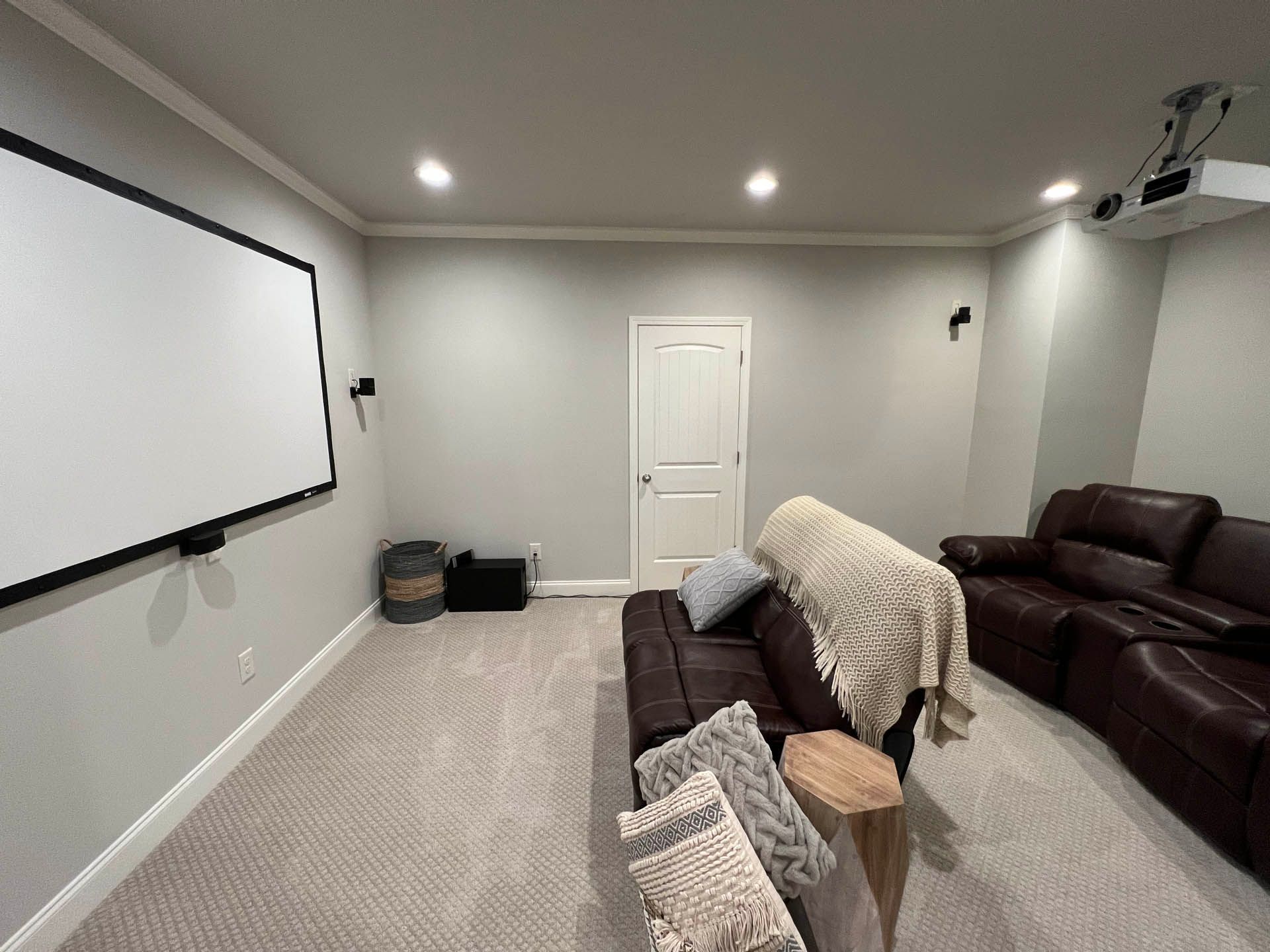A home theater room featuring a white projector screen, brown leather couch, and projector. The room has neutral colored walls and carpet.