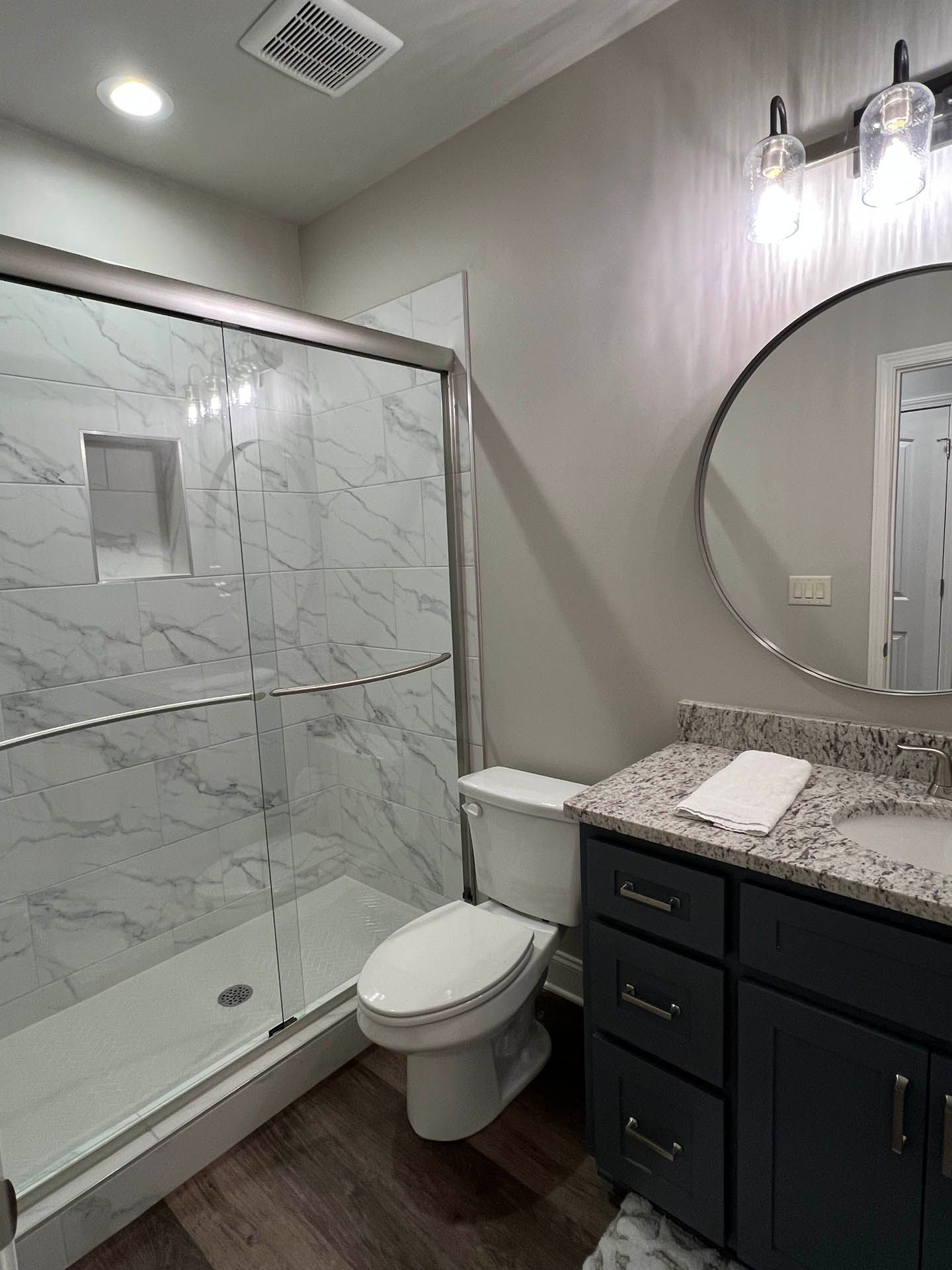 Bathroom with a marble-tiled shower, dark blue vanity, round mirror, and a toilet. The walls are gray.