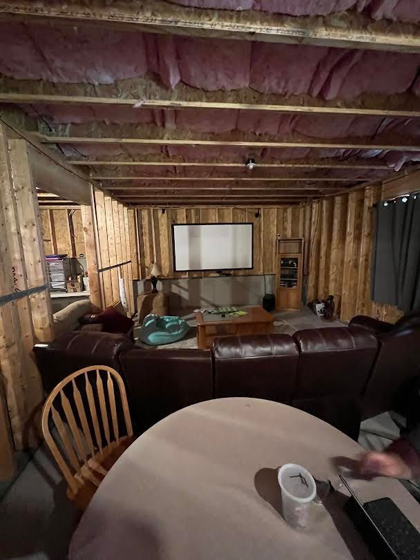 A finished basement living space. Brown leather couch, wooden paneling, and a projector screen. A round table and chair are in the foreground.