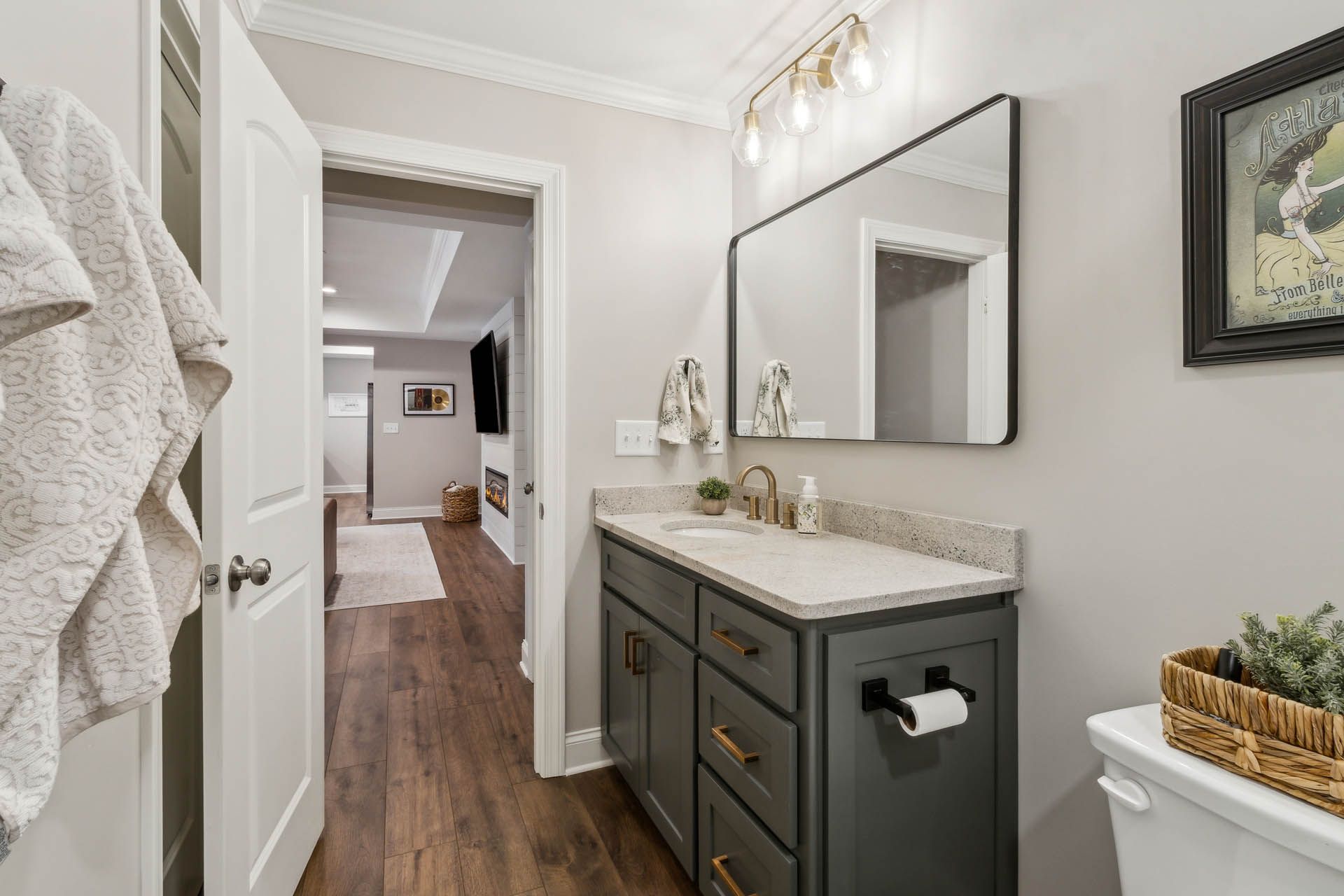 Bathroom with a gray vanity, rectangular mirror, and open door leading to a hallway with a TV and fireplace.