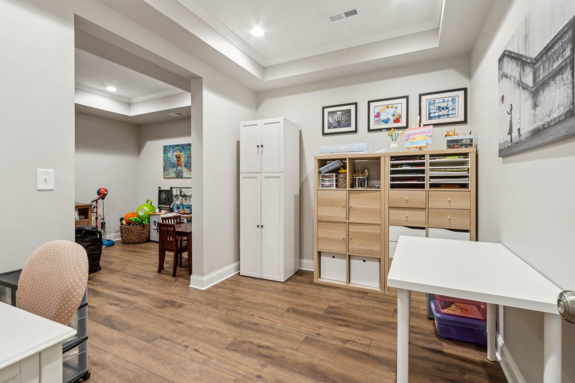 A child's playroom with light-colored walls, hardwood floors, a white storage cabinet, and children's art supplies on a wooden shelf.
