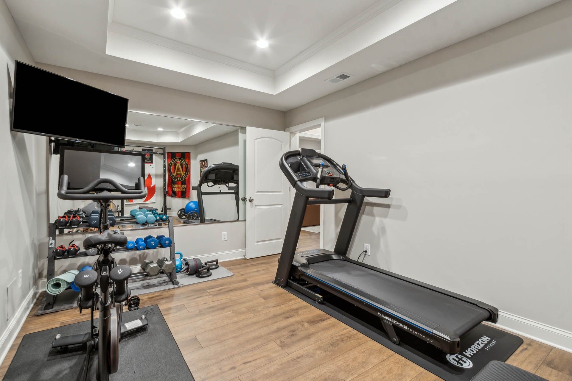 A home gym with a treadmill, stationary bike, weights, and a TV. Mirror on the wall reflects equipment.