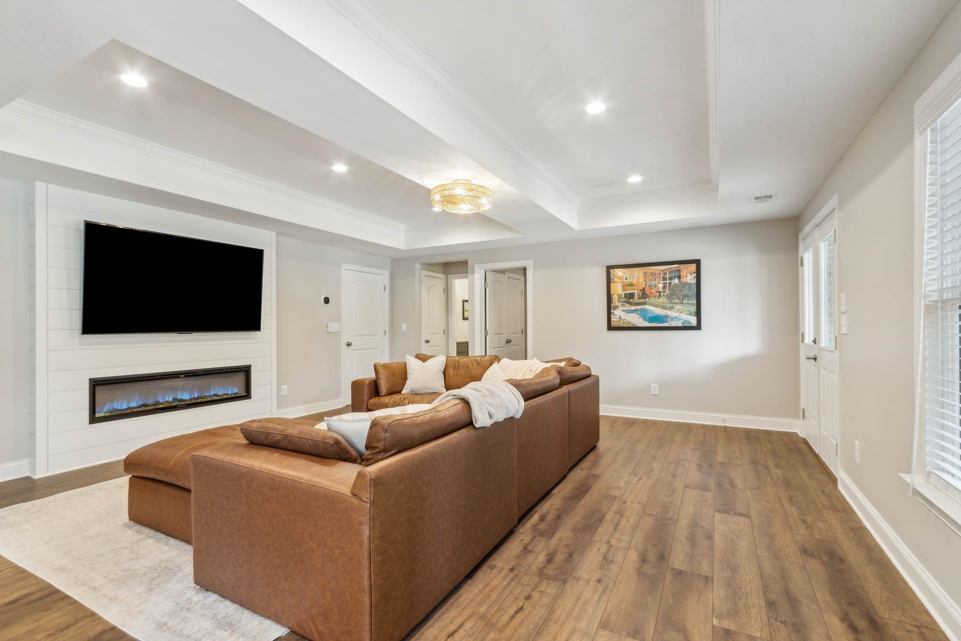 Spacious living room with a brown leather sectional sofa, fireplace, TV, and wood flooring. Soft white walls and recessed lighting.