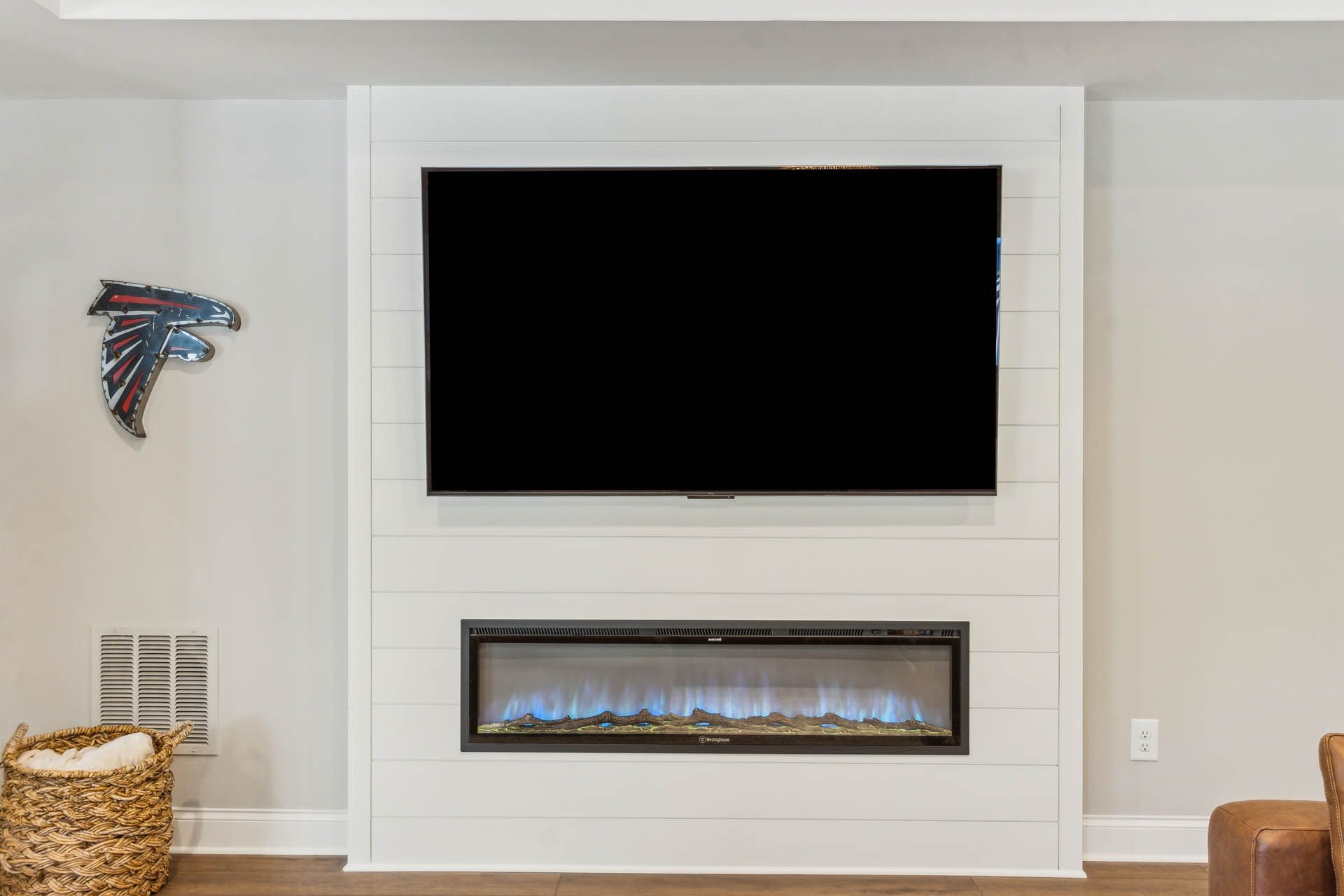 A wall-mounted TV above a modern fireplace with blue flames, set in a white-paneled wall. An Atlanta Falcons emblem is on the left.