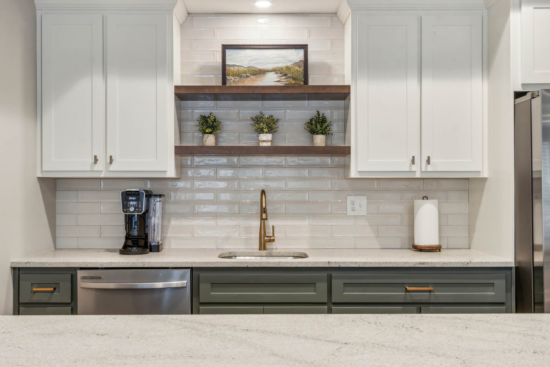 Kitchen with white upper cabinets, green lower cabinets, and a neutral countertop. A sink is centered below a tiled backsplash and wooden shelves.