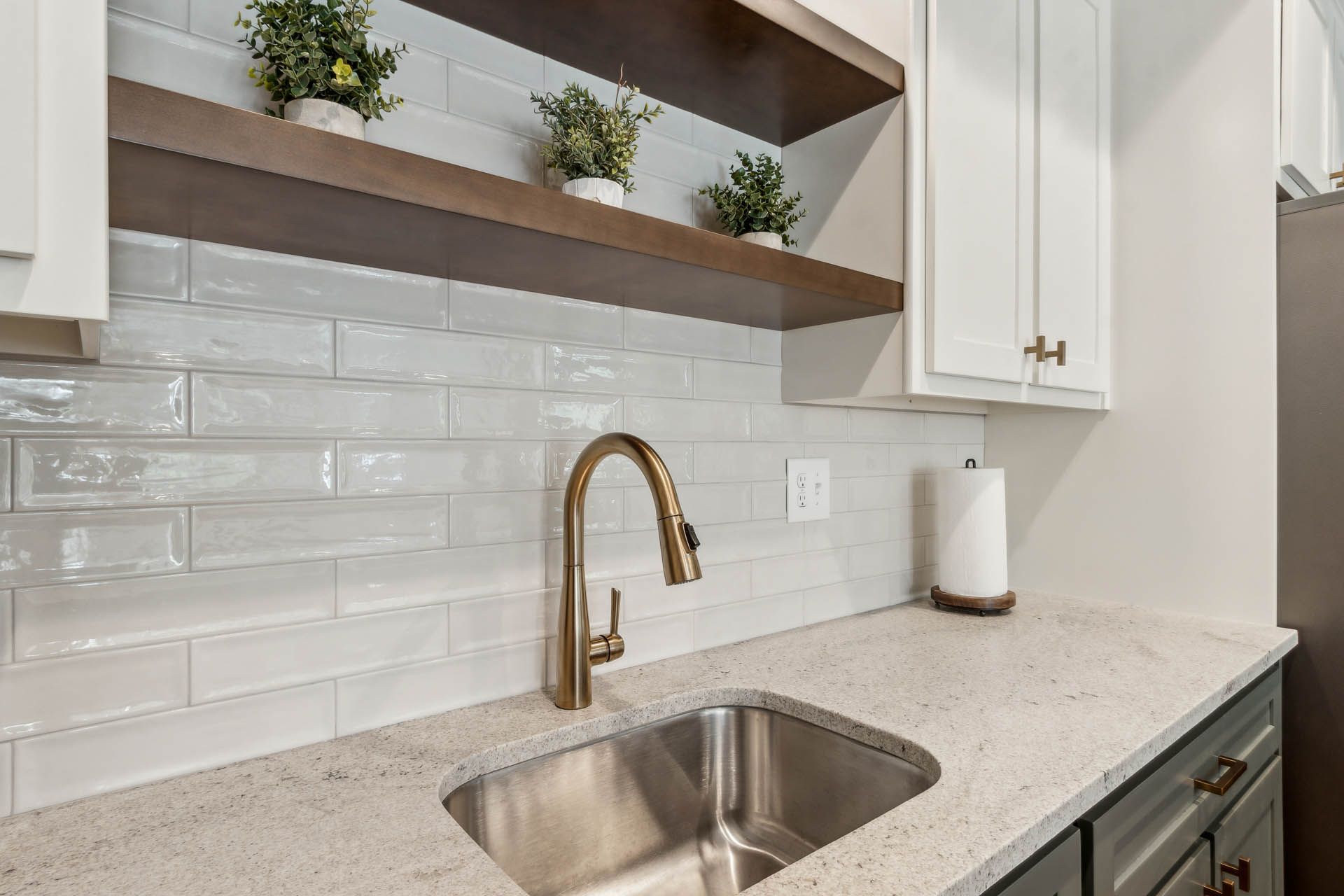 Kitchen with white cabinets, light-colored countertop, stainless steel sink, gold faucet, and white backsplash. Wooden shelves hold plants.