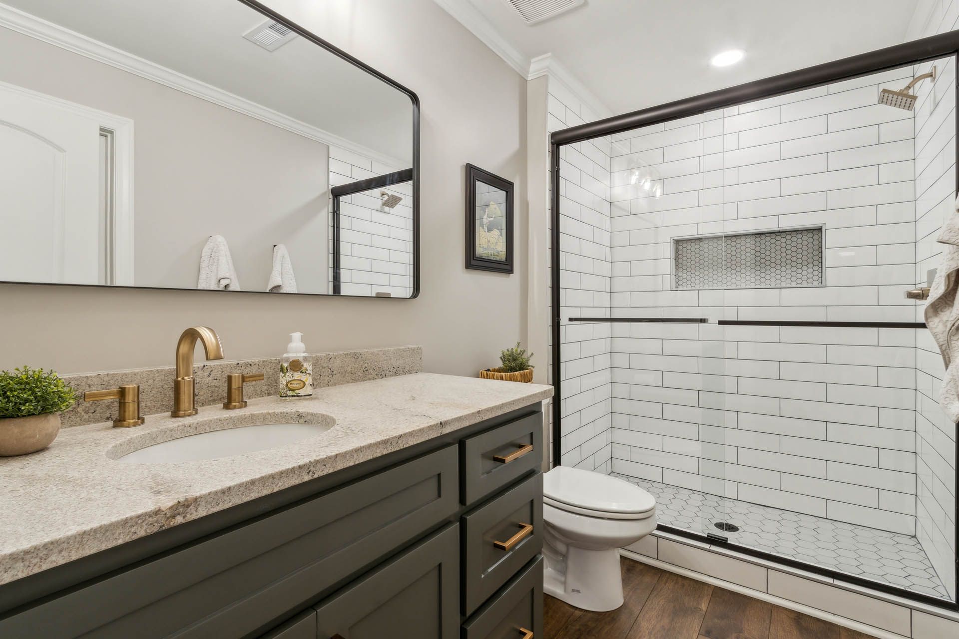 Bathroom with dark gray vanity, gold fixtures, white subway tile shower, and a large black-framed mirror.