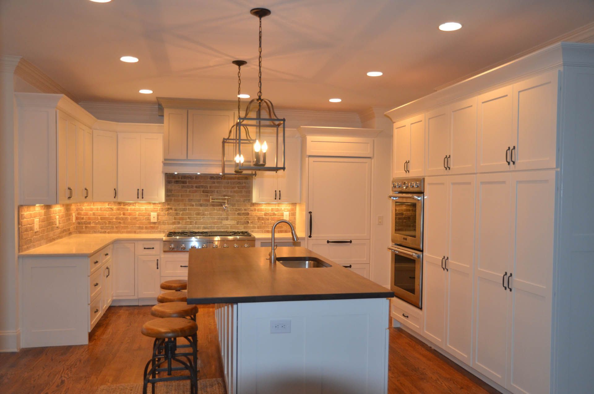 White kitchen with island, cabinets, and stone backsplash. Recessed and pendant lights illuminate the space with bar stools at the island.