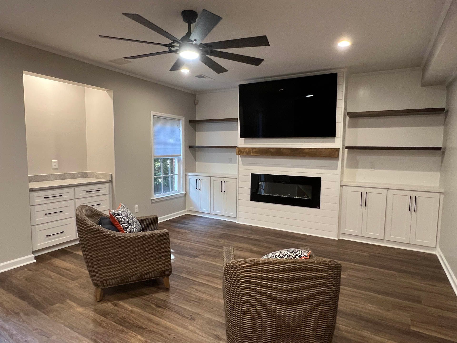 A modern living room with built-in shelves, a fireplace, and a large TV. Two wicker chairs sit on the wooden floor.
