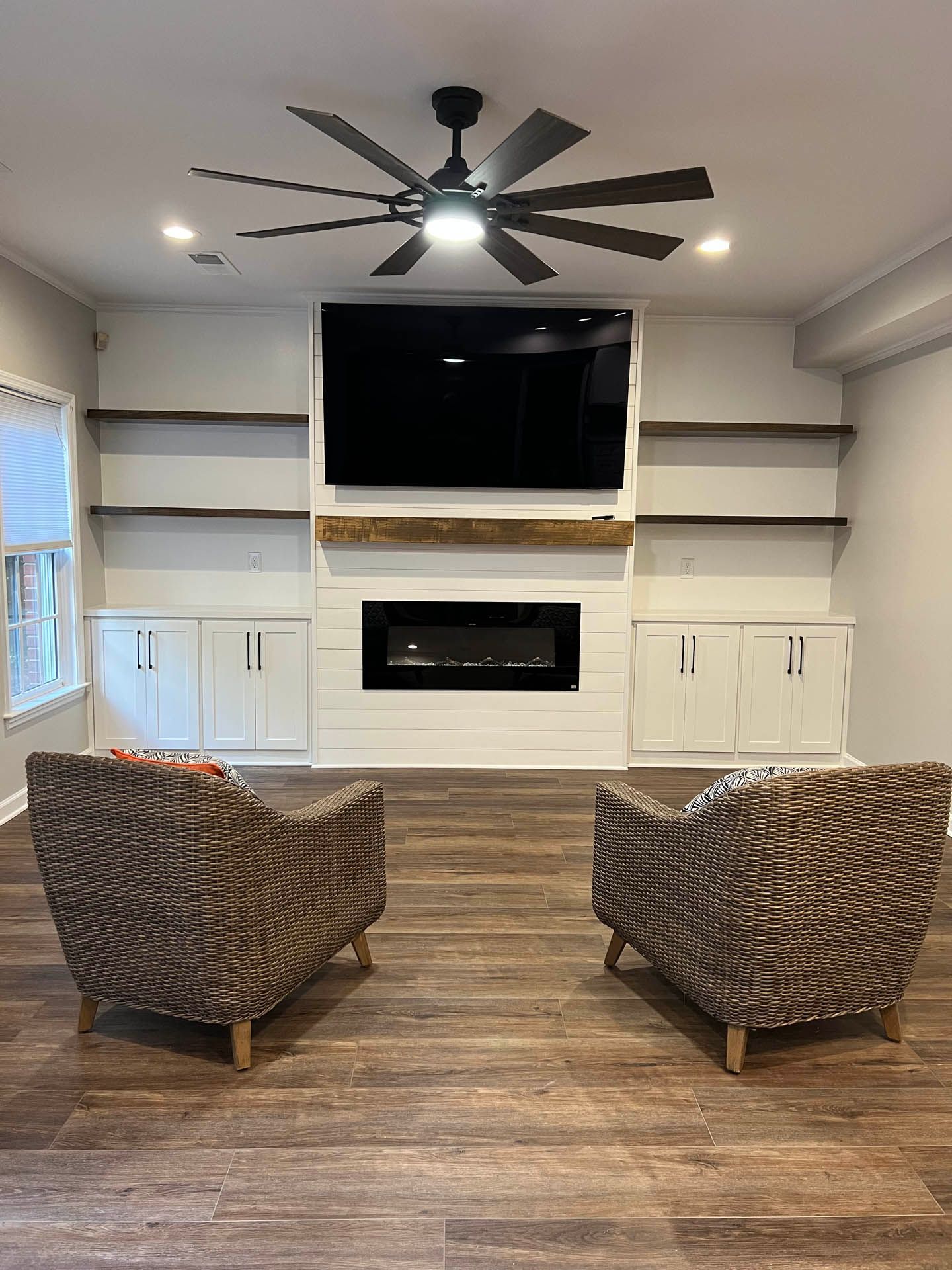 Living room with two patterned armchairs facing a fireplace and large TV.  Dark wood floor, white built-ins, and a ceiling fan.