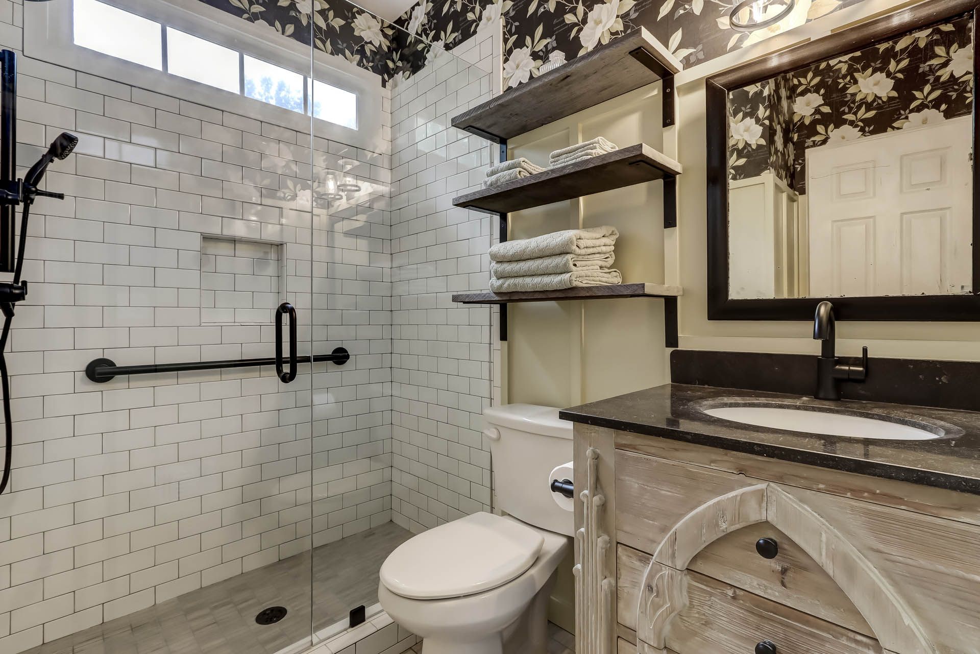 A bathroom featuring white tile, a glass shower, a toilet, a vanity, and dark wood shelves. Black and white tree design wallpaper covers a portion of the wall.
