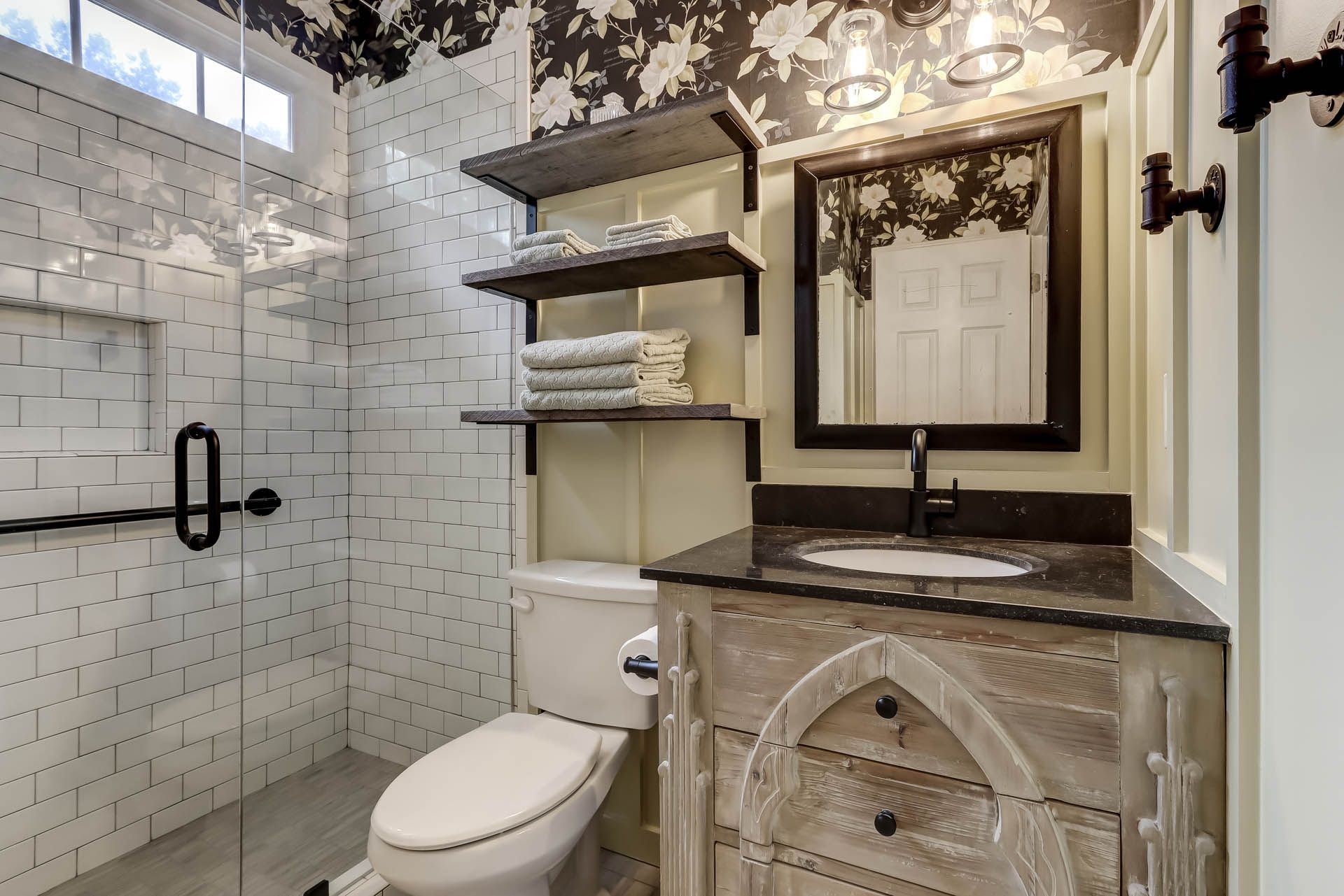 Small bathroom with white brick-like tiles, black floral wallpaper, and a vanity with a rustic-style cabinet. A glass shower and open shelves are also visible.
