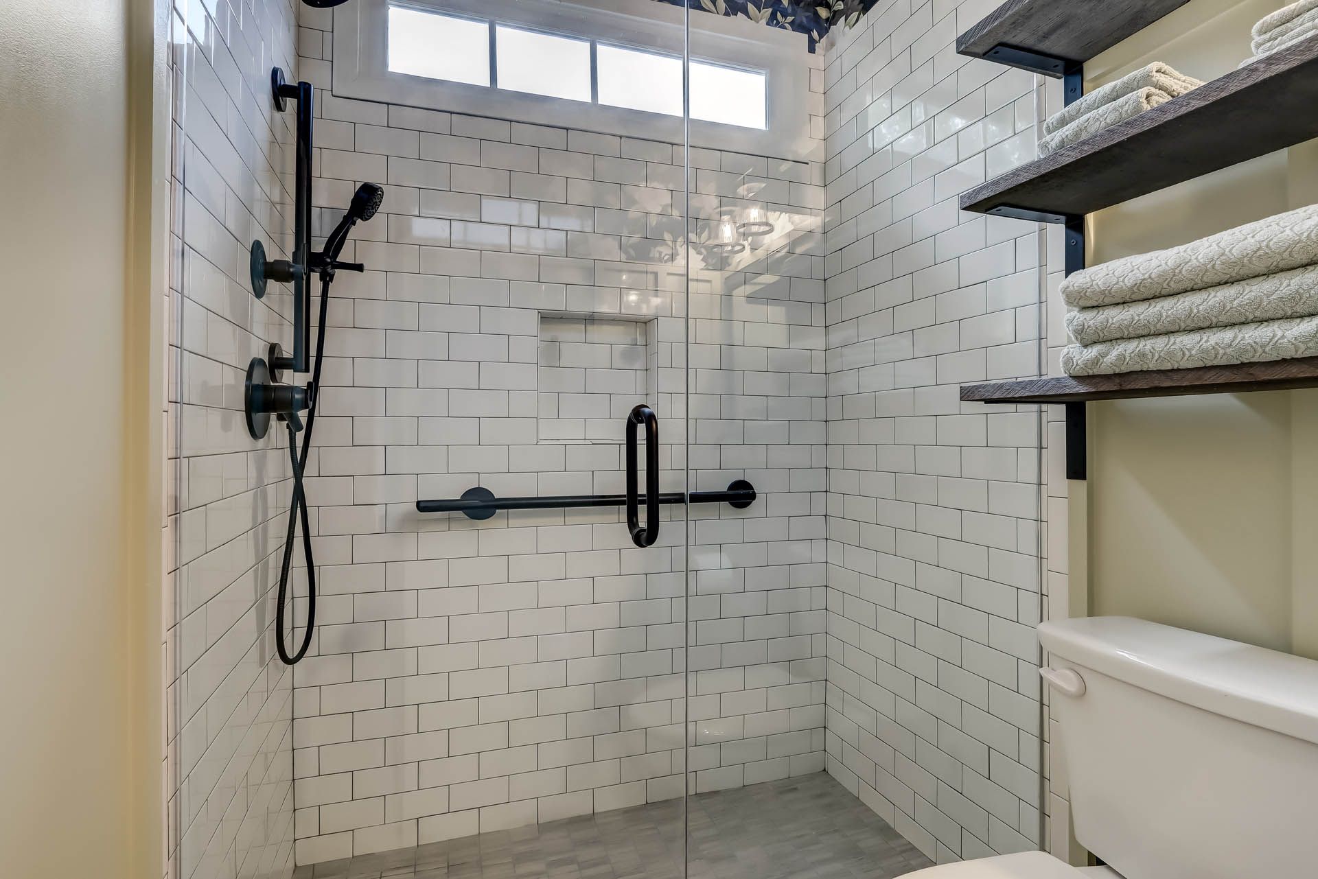 A modern bathroom featuring a glass-enclosed shower with black fixtures, white subway tile, and open wooden shelves with towels.