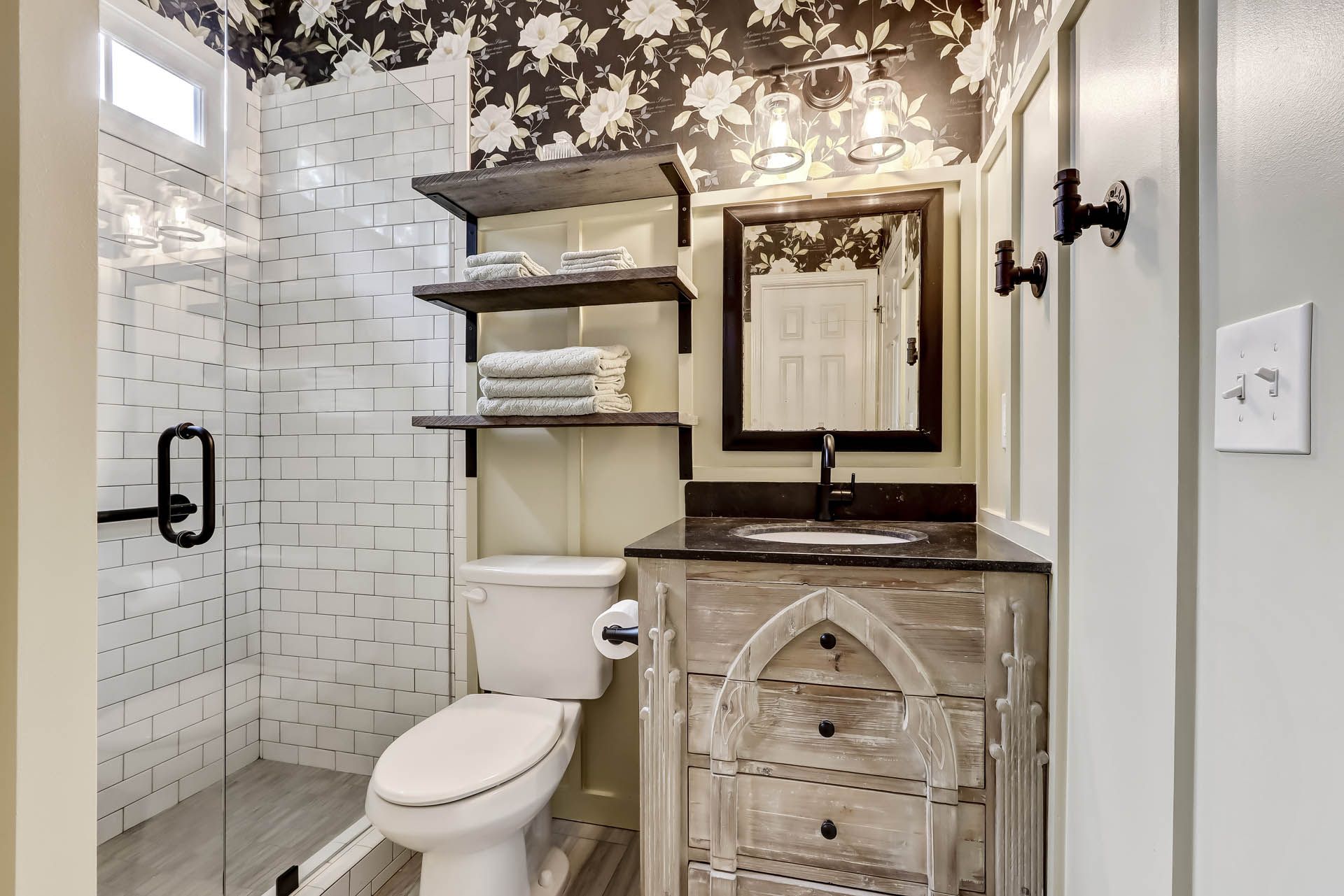 Small bathroom with white tile shower, patterned wallpaper, rustic vanity, and open shelving.