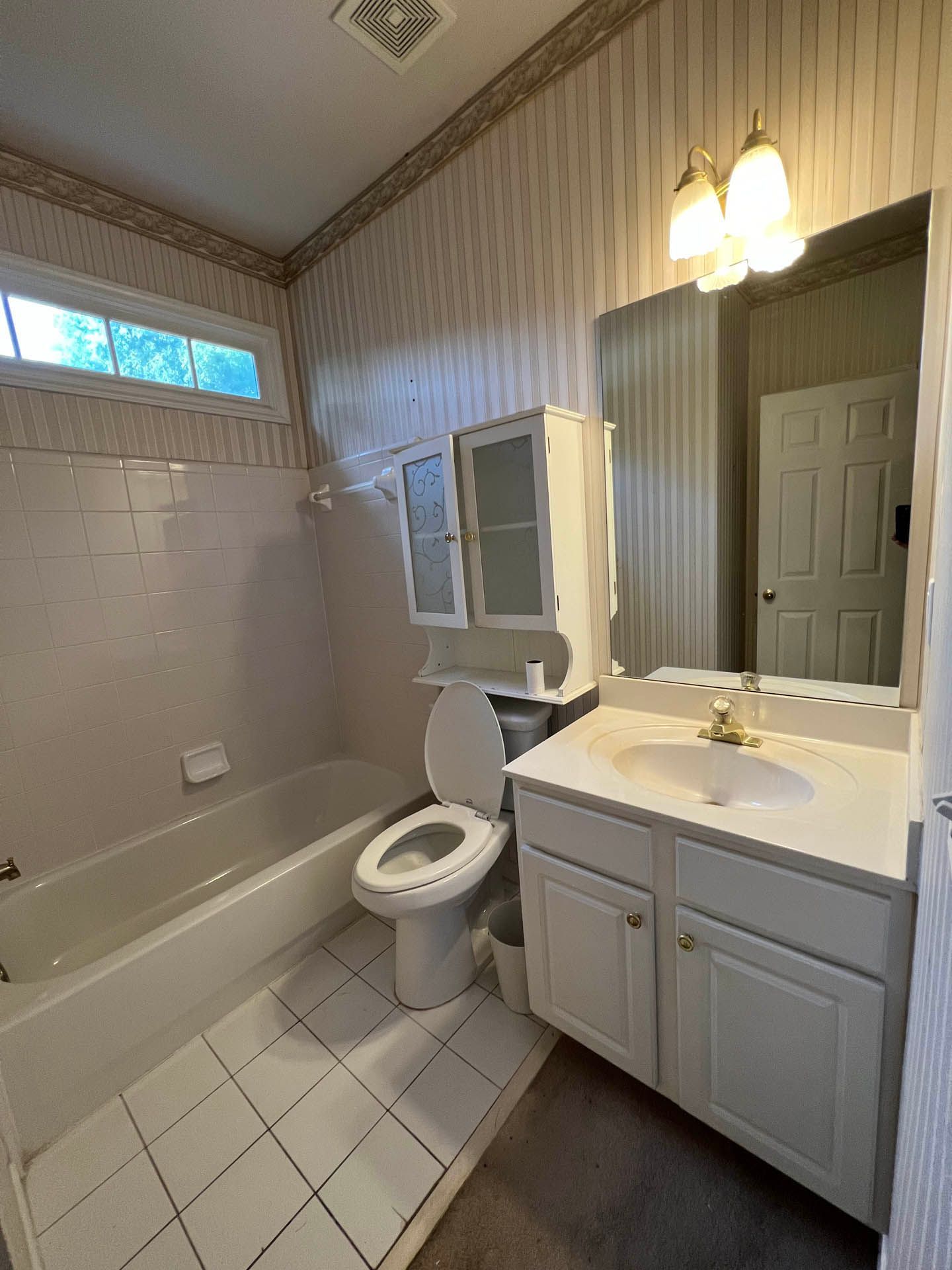 Bathroom with a white tub, toilet, vanity, and overhead cabinet. Beige wallpaper and white tile flooring.