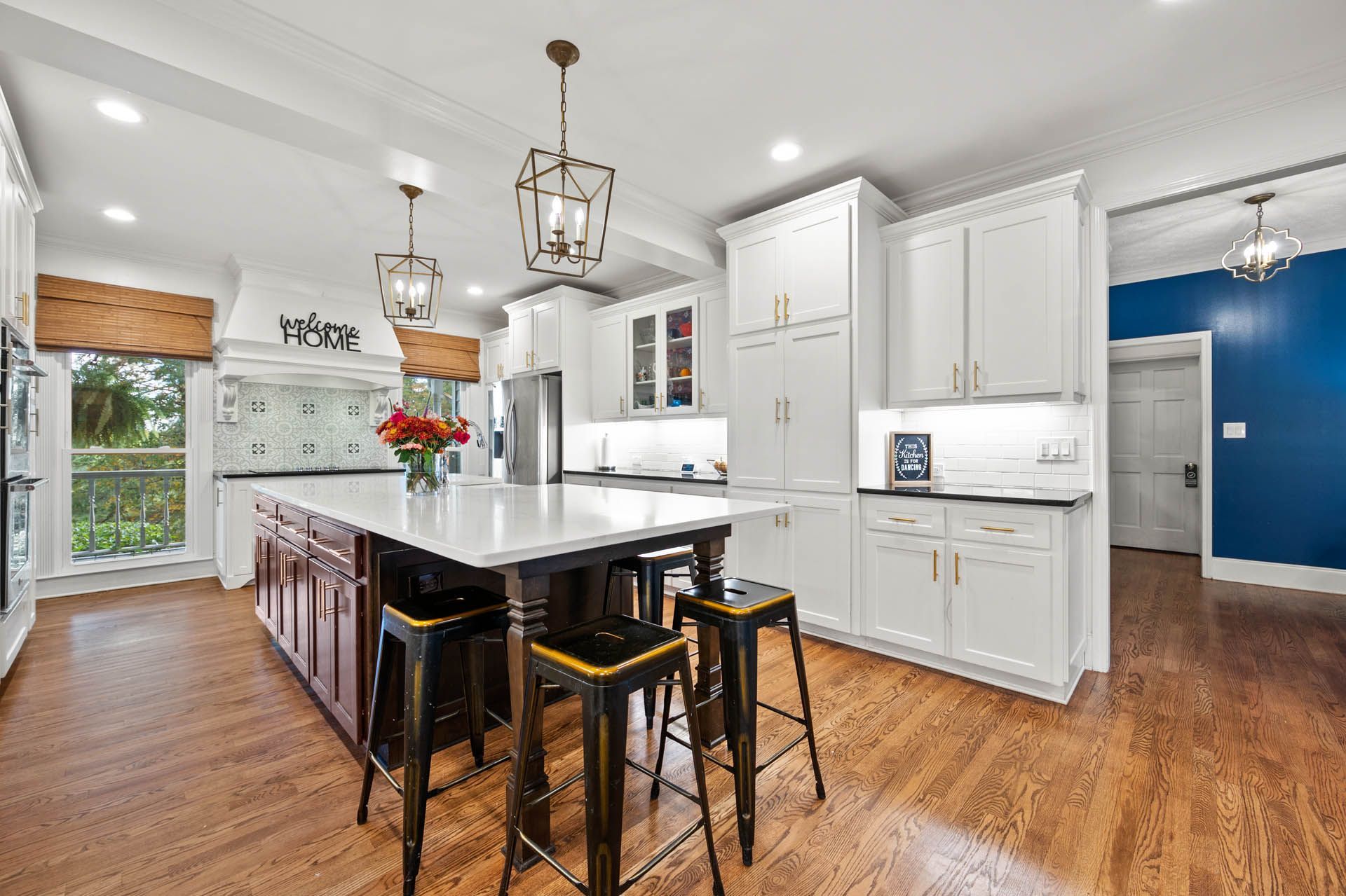 Spacious white kitchen with a large island, dark cabinets, and wooden floors. Black bar stools sit at the island, and a blue doorway is visible in the background.