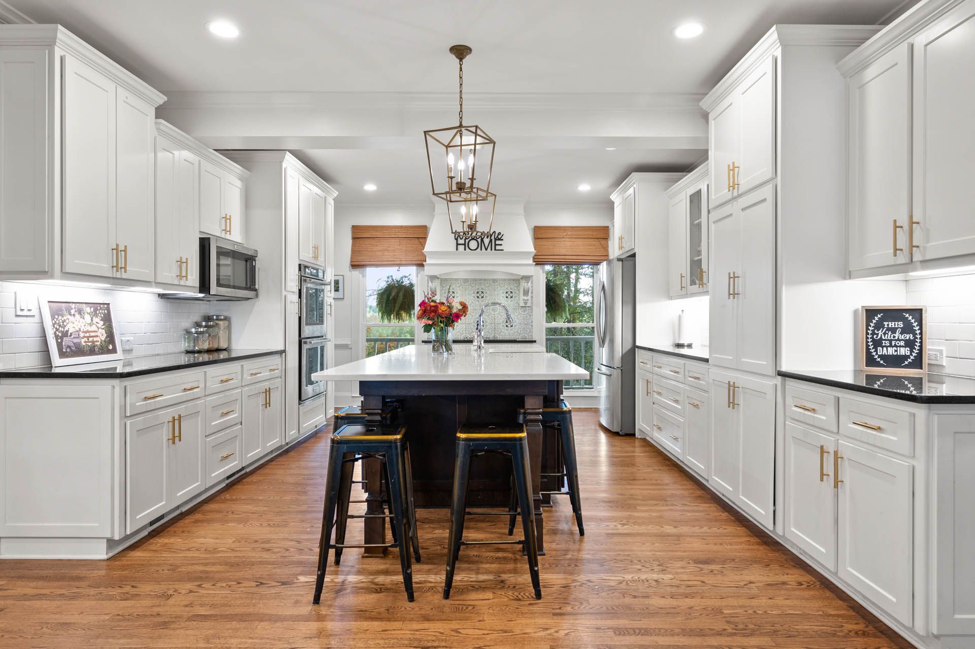 Spacious white kitchen with dark island, three black stools, and wooden flooring. A central chandelier hangs above the island.