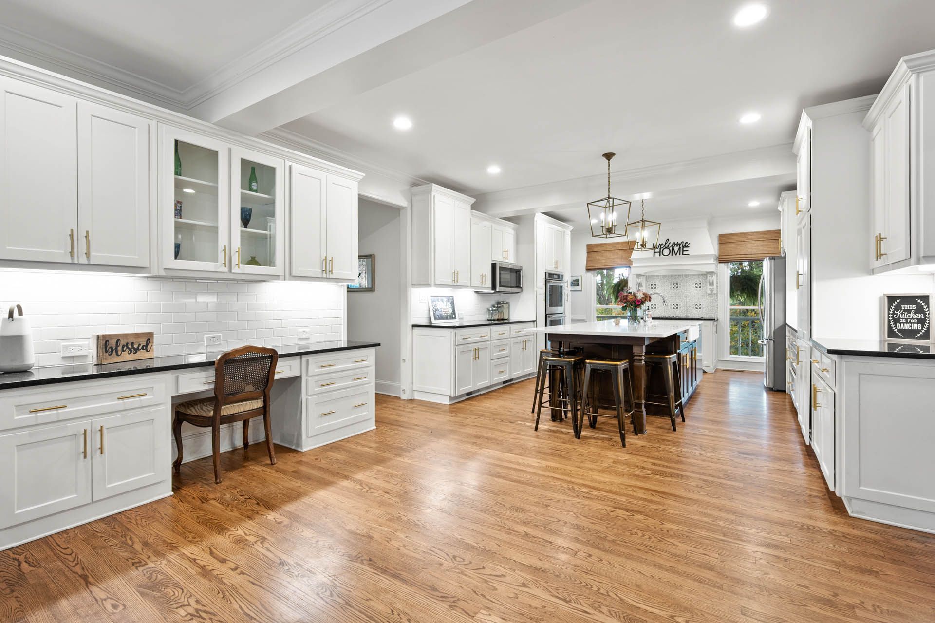 Spacious white kitchen with wood floors, dark island, and built-in desk. Bright, airy room with cabinets and appliances.