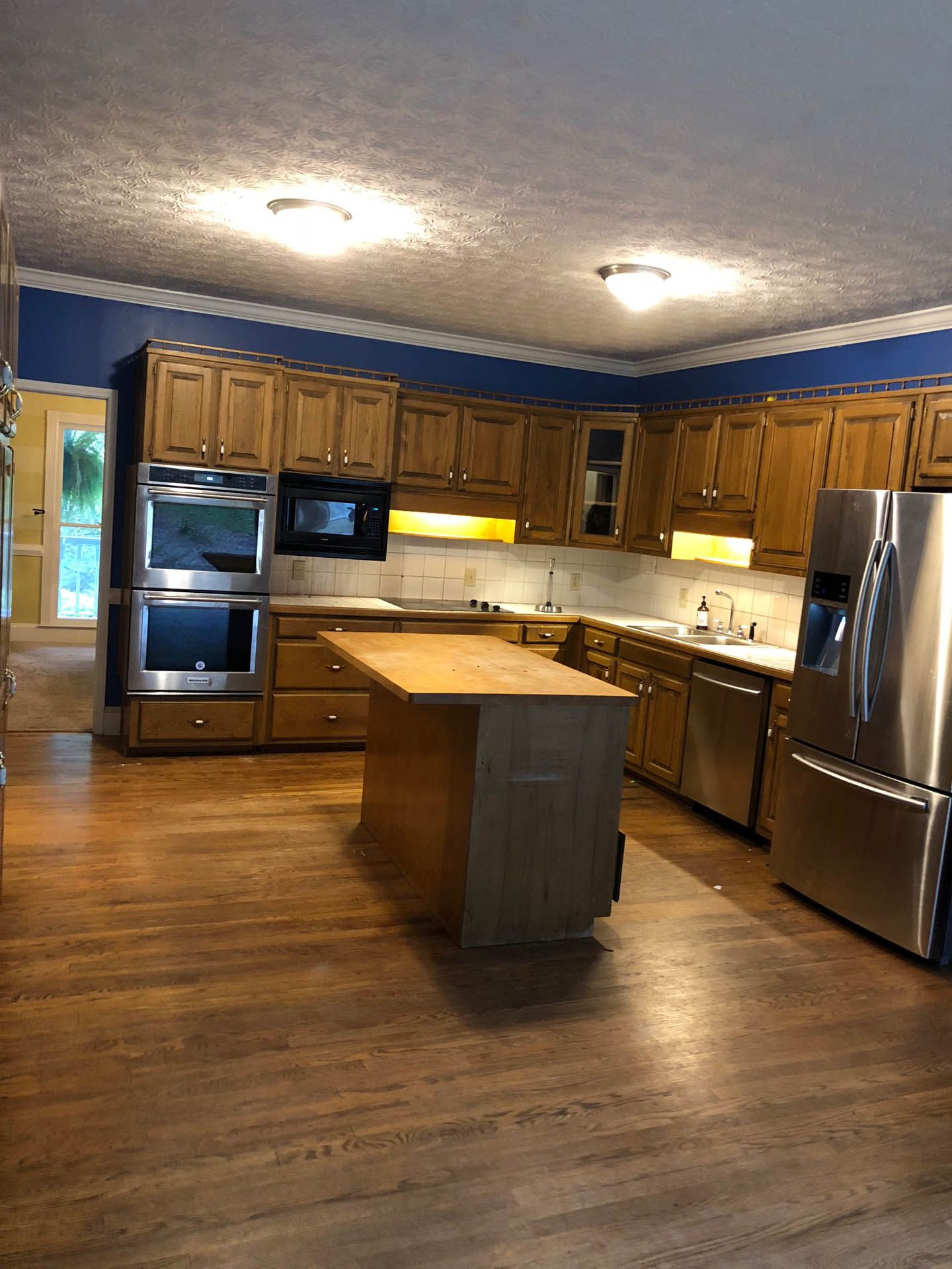 A kitchen with wood cabinets, a stainless steel fridge, and a center island. The room has wooden floors and blue walls.