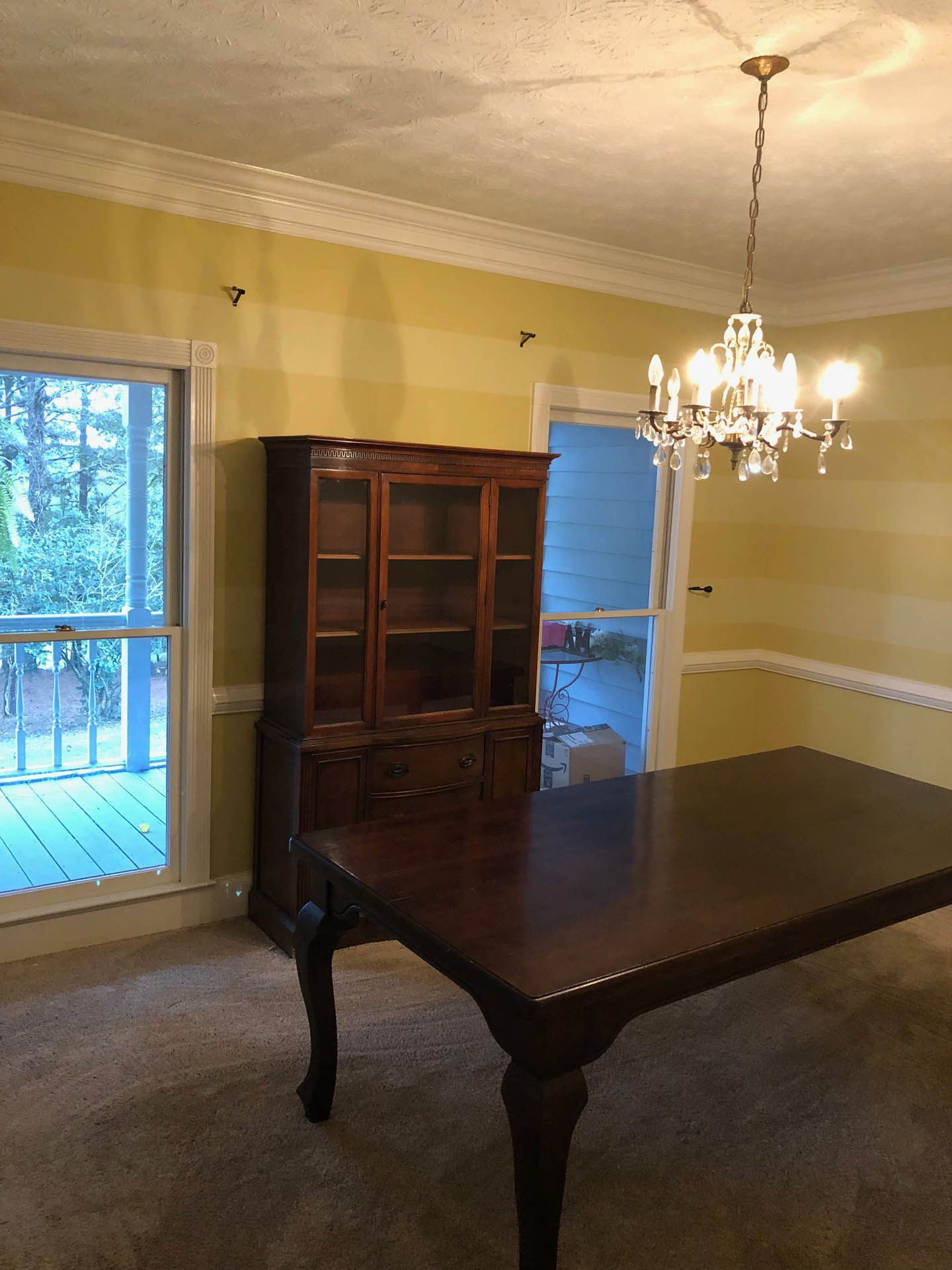 Dining room with wooden furniture, including a table and china cabinet, against yellow striped walls. A chandelier hangs above the table near a window and doorway.