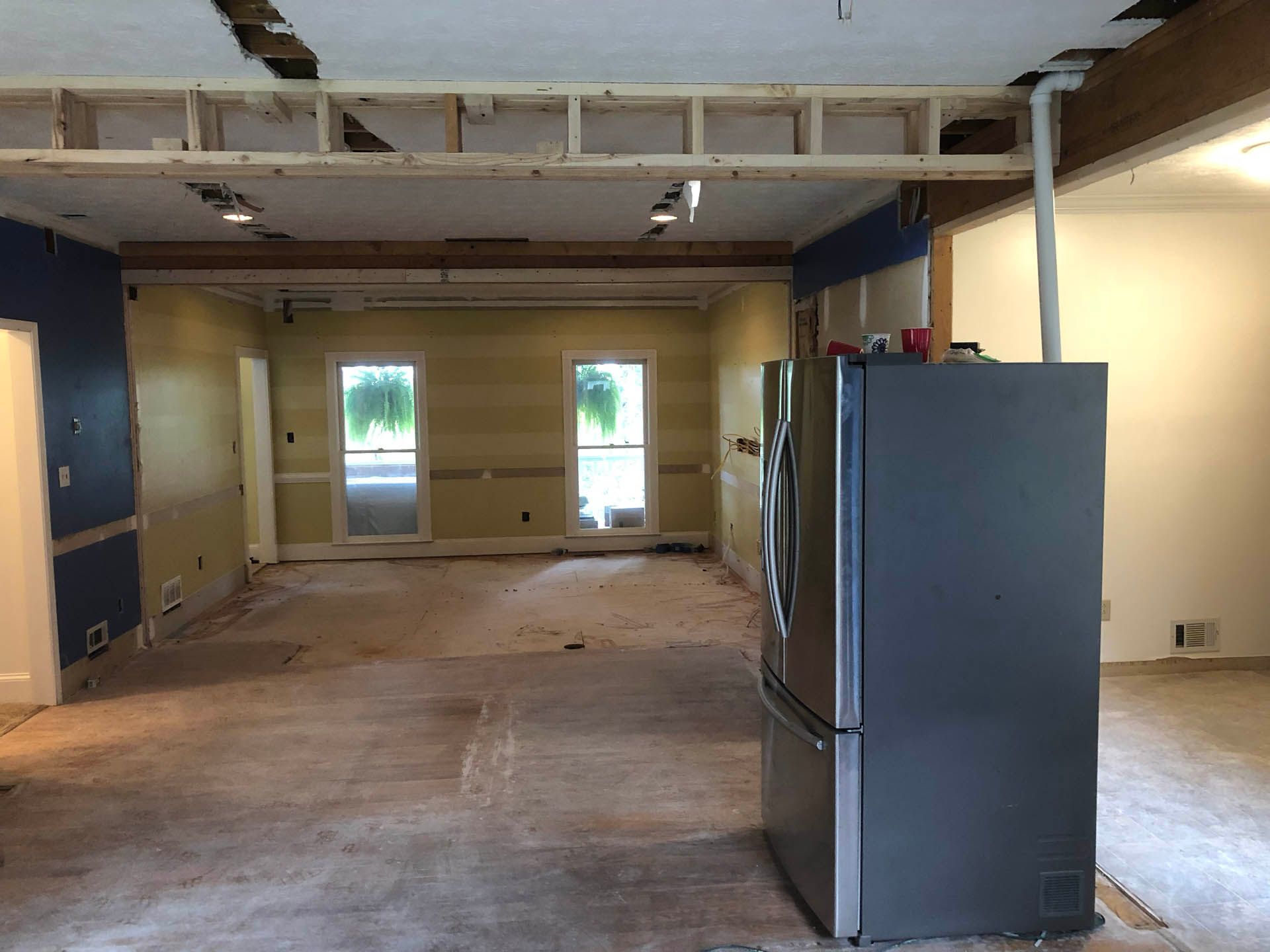 Interior during renovation; unfinished room with exposed ceiling beams, refrigerator, and walls in various stages of completion.