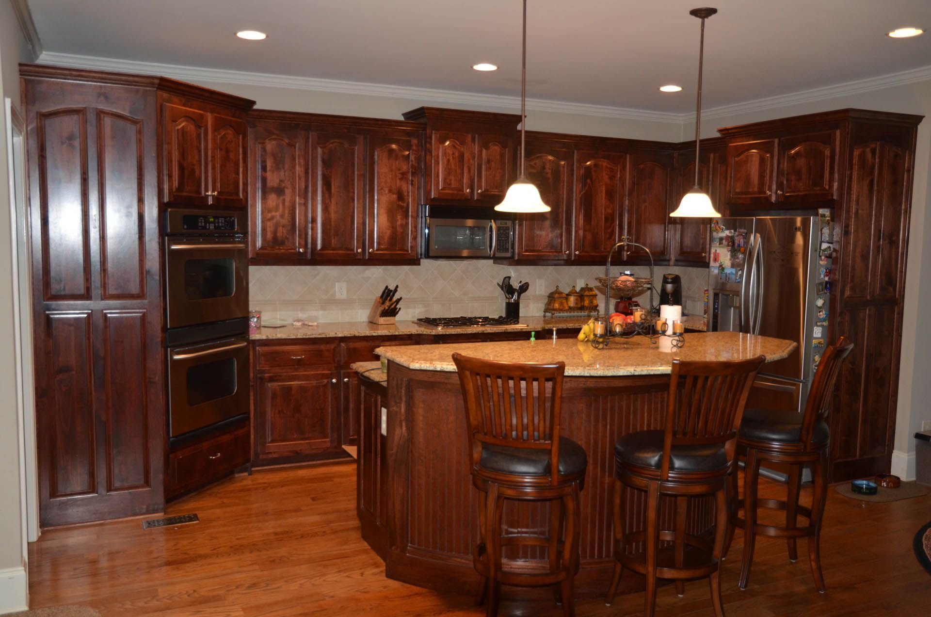 A kitchen with dark wood cabinets, a curved island with bar stools, and light-colored countertops. The kitchen has stainless steel appliances and recessed lighting.