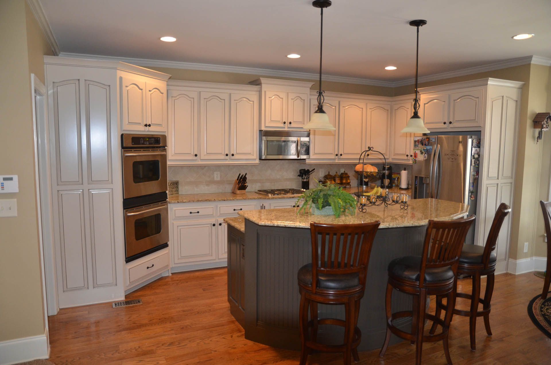 Creamy white kitchen with island, hardwood floors, and stainless steel appliances. Two pendant lights hang above the island with barstools.