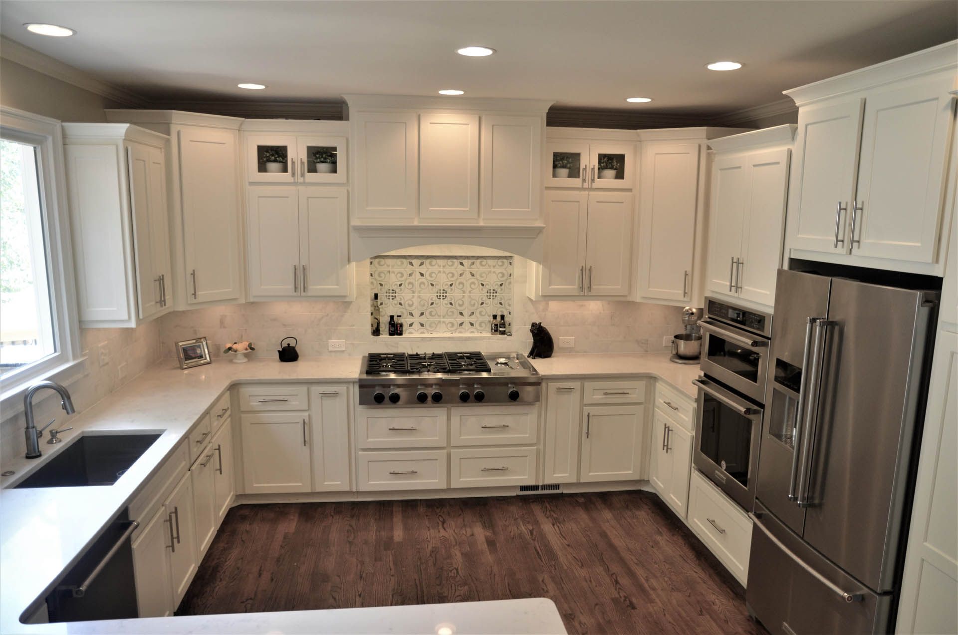 White kitchen with light-colored countertops, stainless steel appliances, and wooden floors. The design includes a cooktop, range hood, and cabinetry.