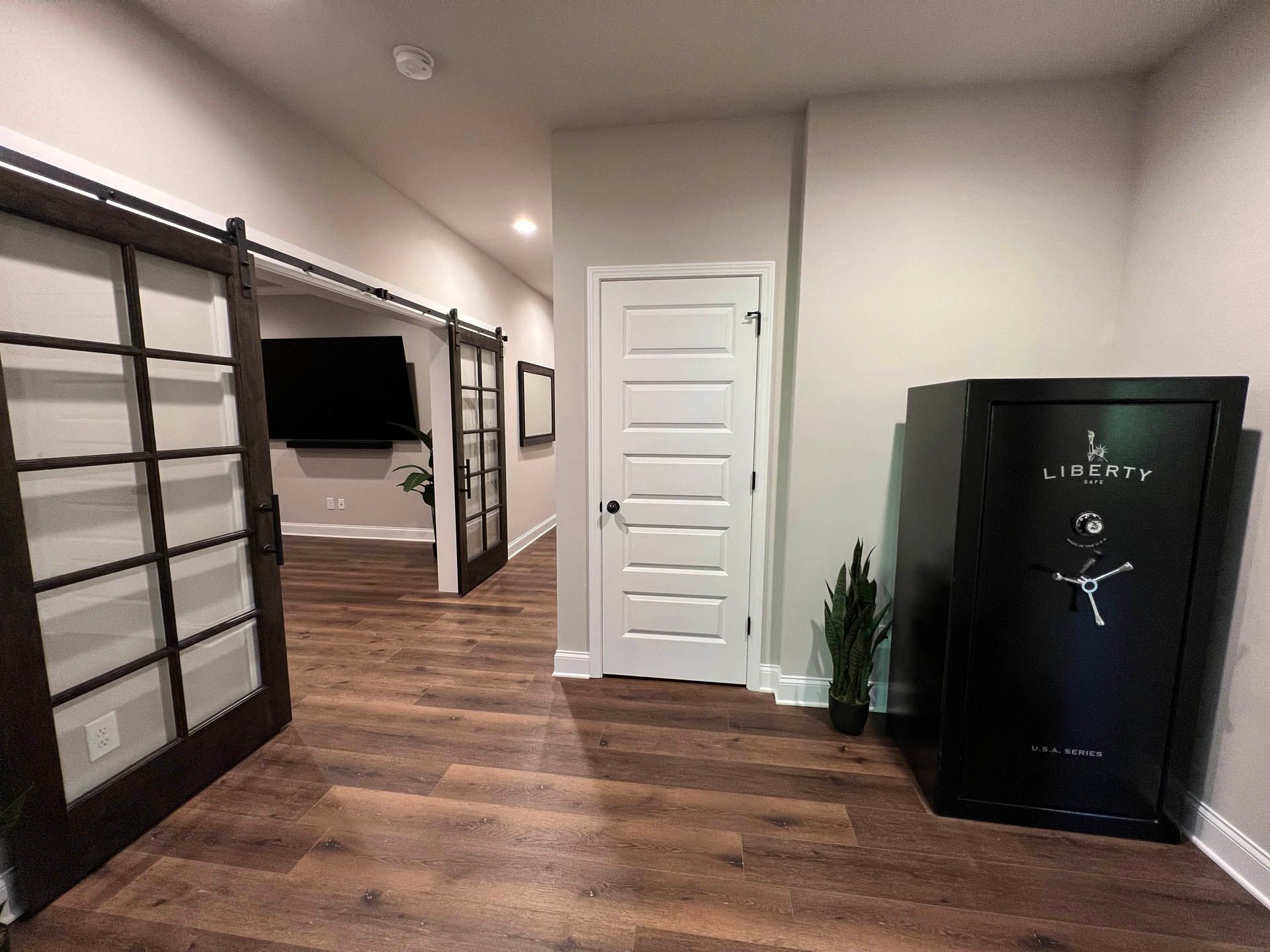 A dark-paneled sliding door opens to a long hallway with a wood floor, leading to another doorway. A gun safe stands to the right.