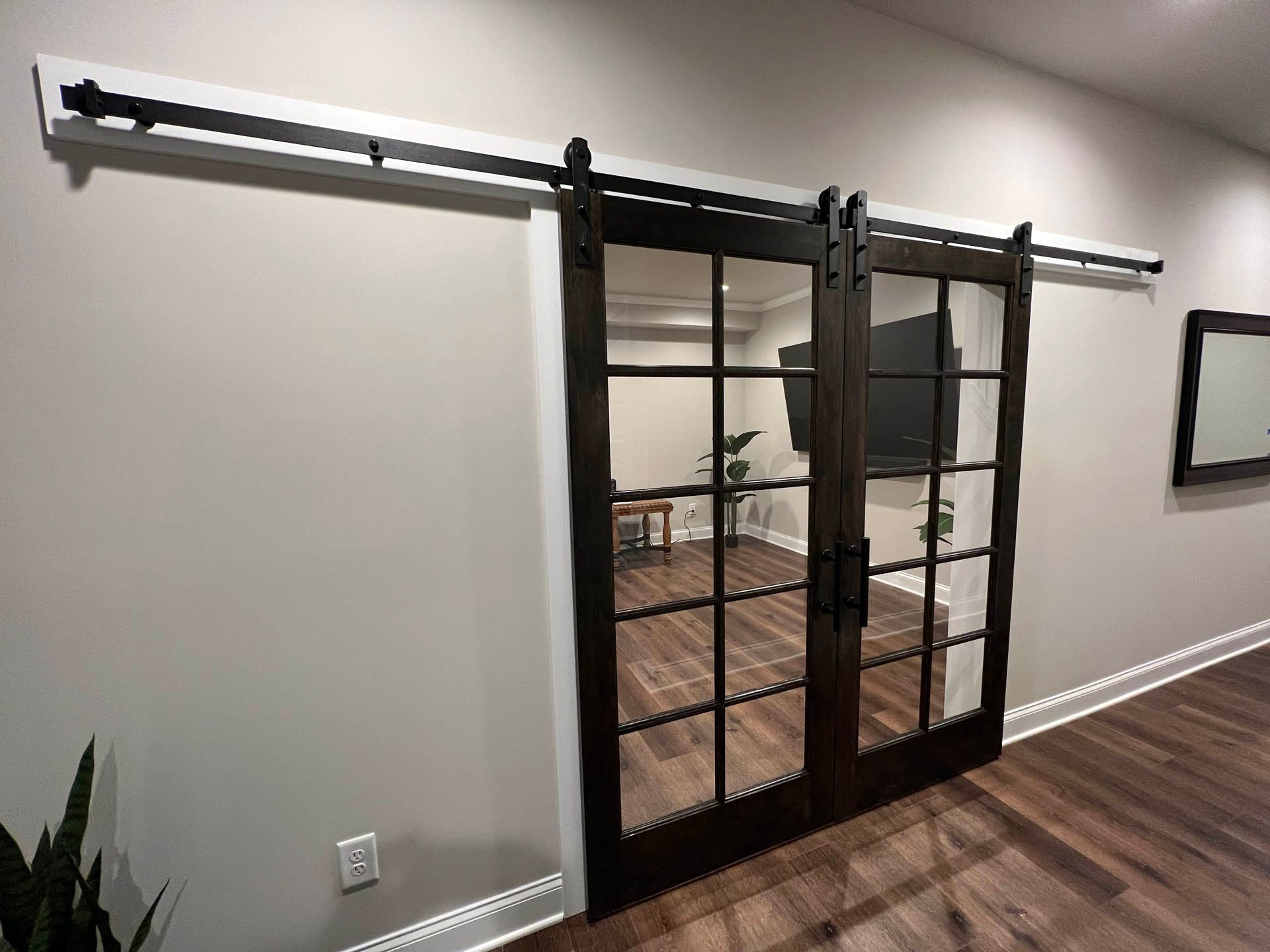 Barn doors with glass panels, dark brown frame, and black hardware, mounted on a white header rail against a gray wall, leading to a room with a TV and hardwood floor.