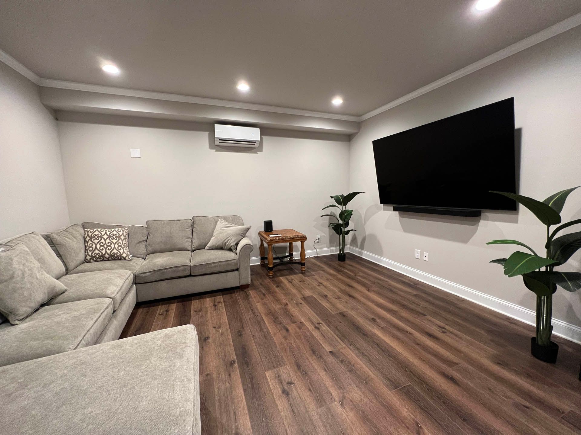 A modern living room with a gray sectional sofa, large TV on the wall, and dark wood flooring. Two potted plants add greenery.