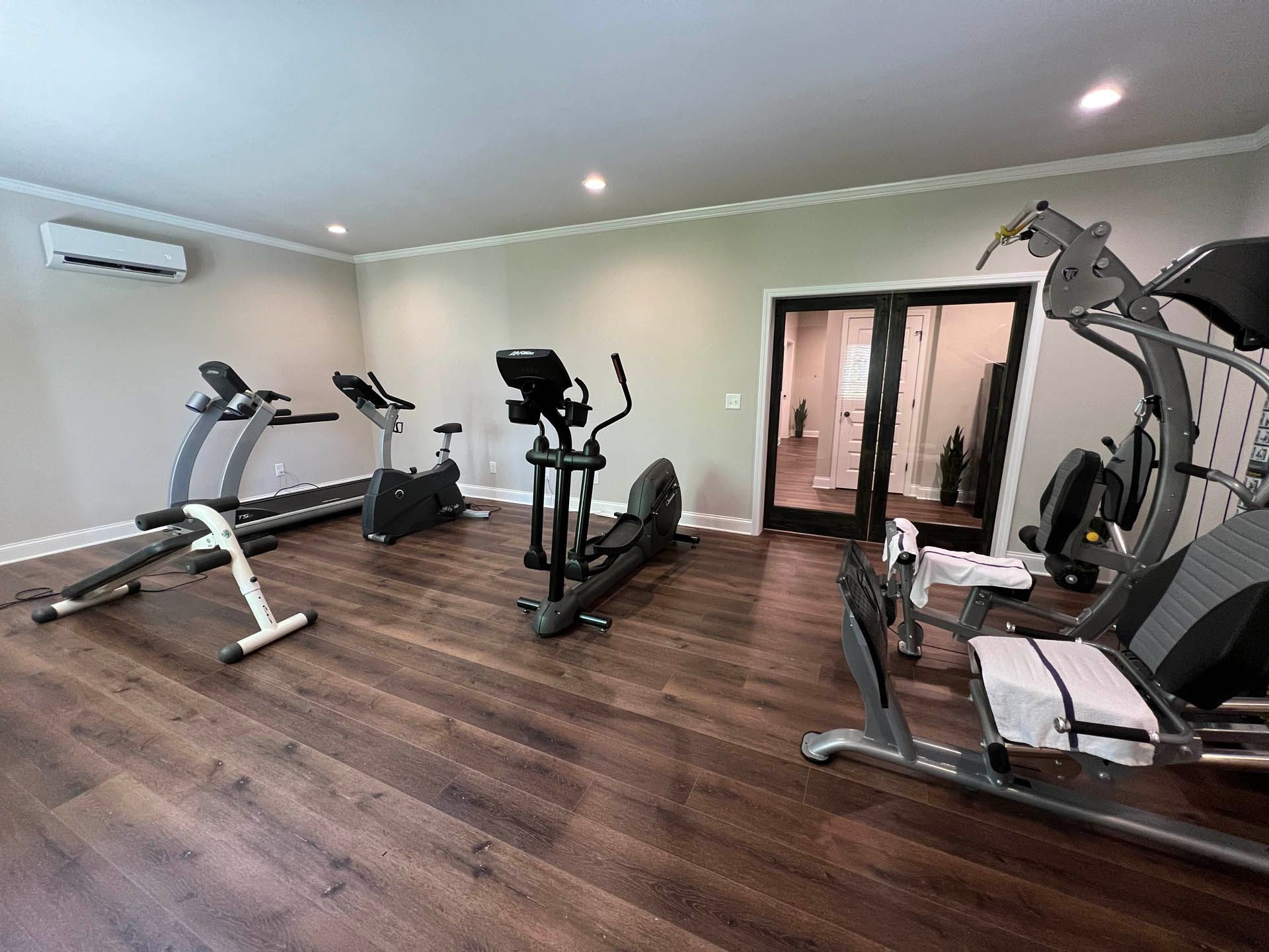 A home gym with several exercise machines on a dark wood floor. Equipment includes a treadmill, stationary bike, elliptical, and a seated elliptical trainer.