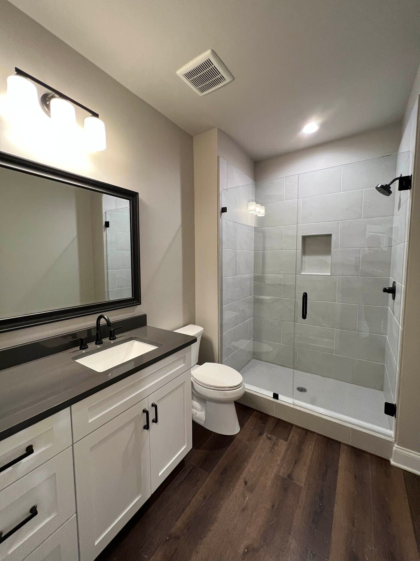 Bathroom with a white vanity, dark countertop, and a glass-enclosed shower with gray tiles. Dark wood-look flooring.
