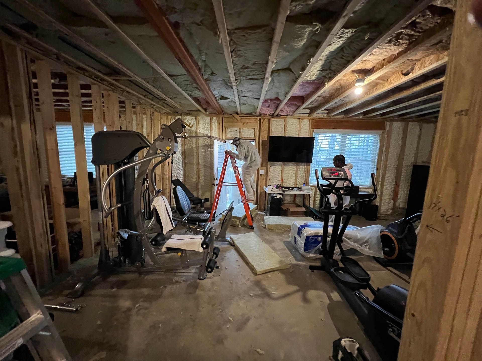 A basement being renovated into a home gym. Exercise equipment sits amidst exposed framing, insulation, and a ladder.