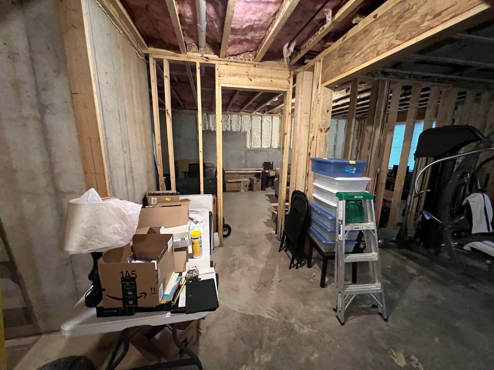 Interior of unfinished basement with exposed wooden framing, concrete walls, and various items, including boxes, a ladder, and storage bins.