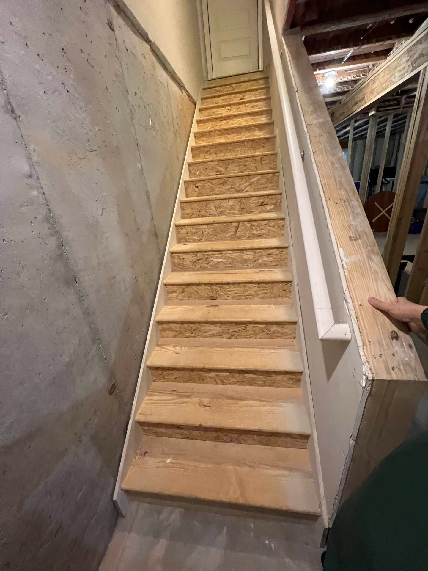 Staircase leading upwards, made of unpainted wood. One side is against a concrete wall, the other has a white handrail.