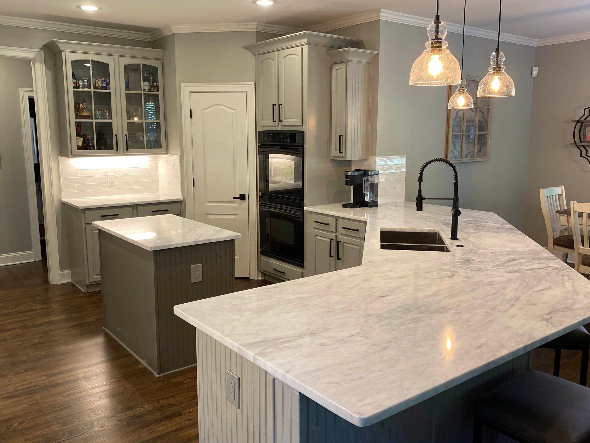 A modern kitchen with gray cabinets, white countertops, and a kitchen island. Pendant lights hang over the island with a black faucet and sink.