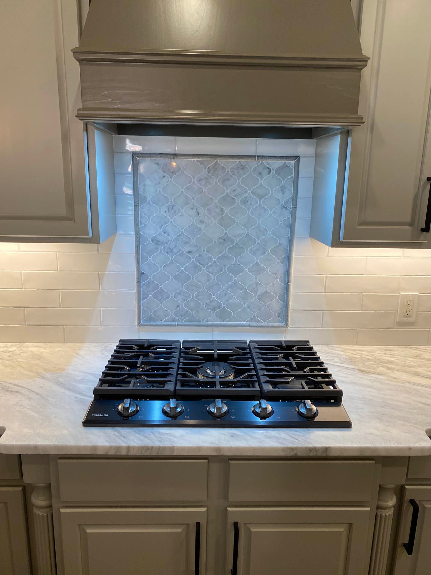 A gas cooktop in a kitchen with a gray range hood, light countertops, and decorative backsplash tiles. Gray cabinets and white subway tiles are also visible.