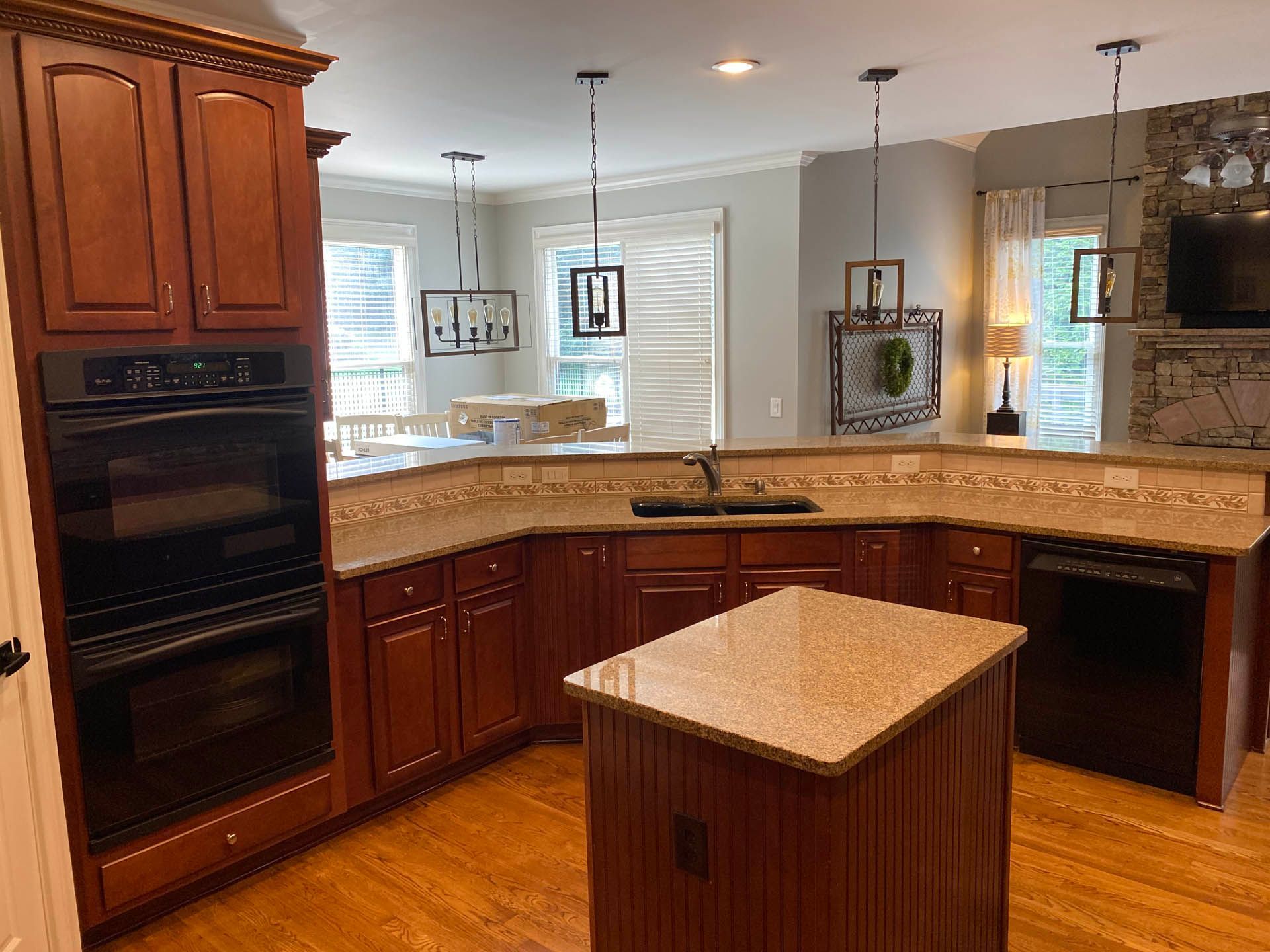 A kitchen with dark wood cabinets, granite countertops, and a central island. There's an oven, sink, and pendant lights in the spacious room.
