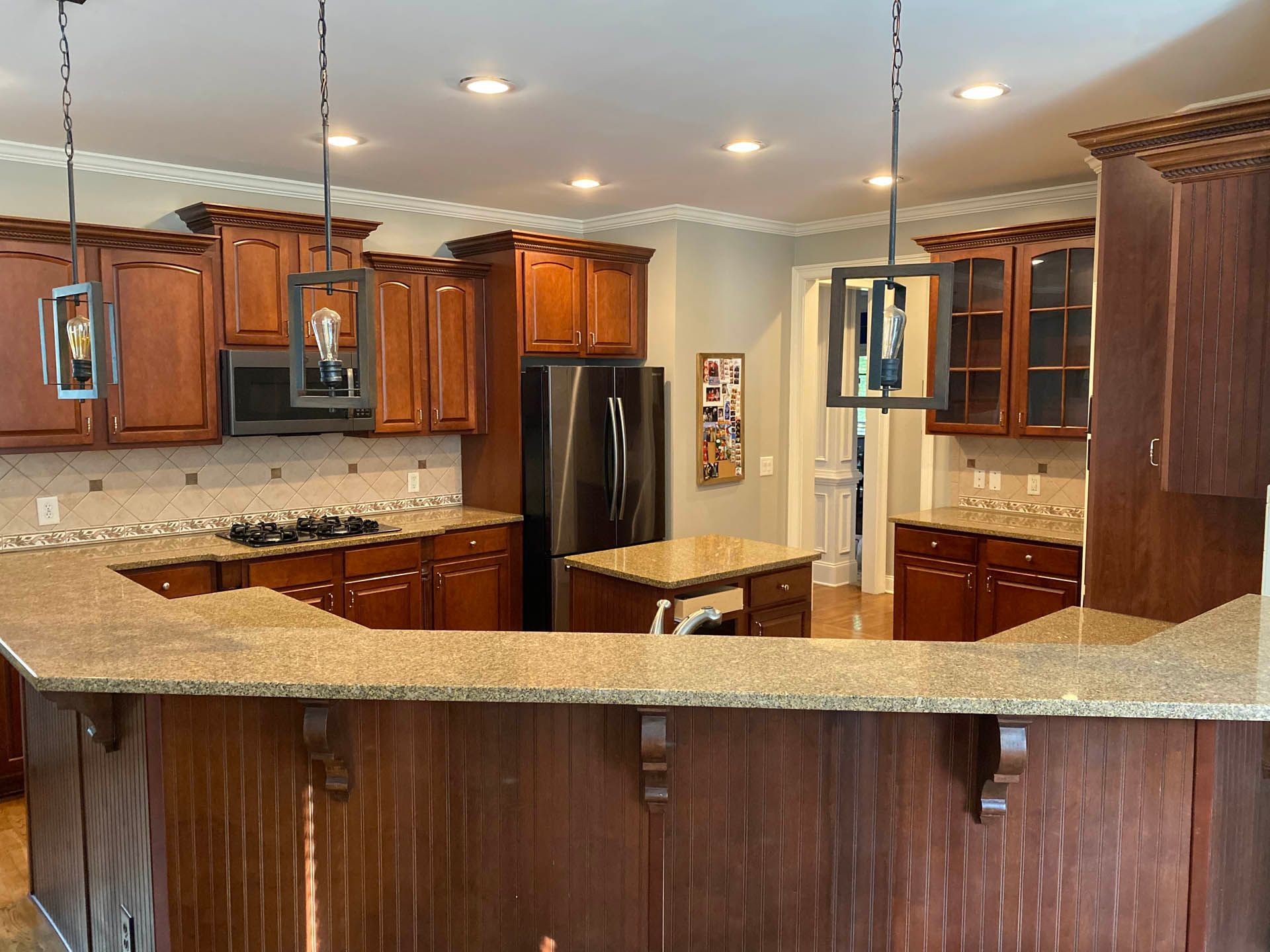 A U-shaped kitchen with brown cabinets, granite countertops, and a stainless steel refrigerator.  Three pendant lights hang above the island.