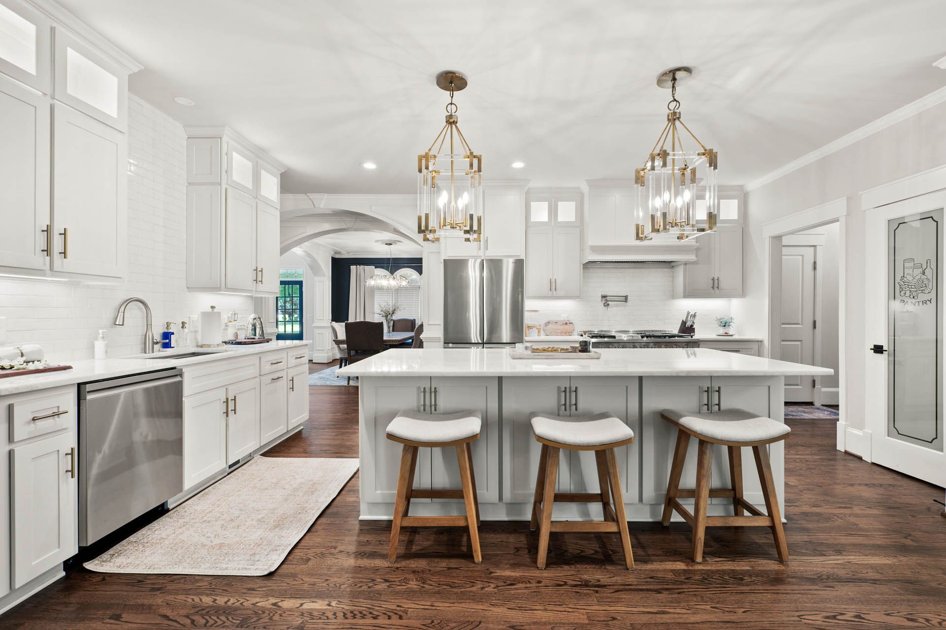 Bright white kitchen with a large island and wooden stools. Dark wood floors, stainless steel appliances, and gold light fixtures.
