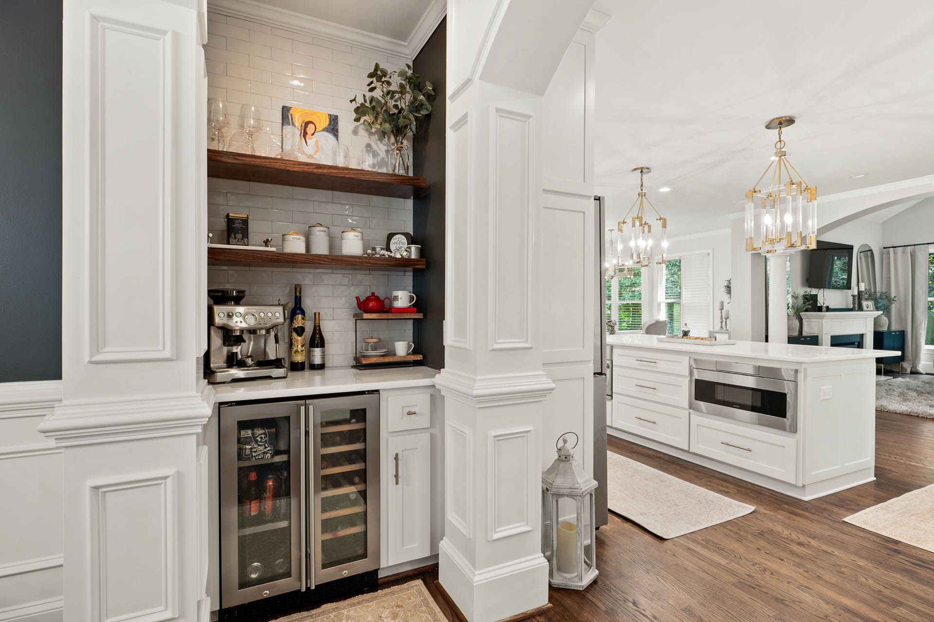 A built-in coffee and wine bar with white cabinetry and open shelving, leading into a bright kitchen with an island.
