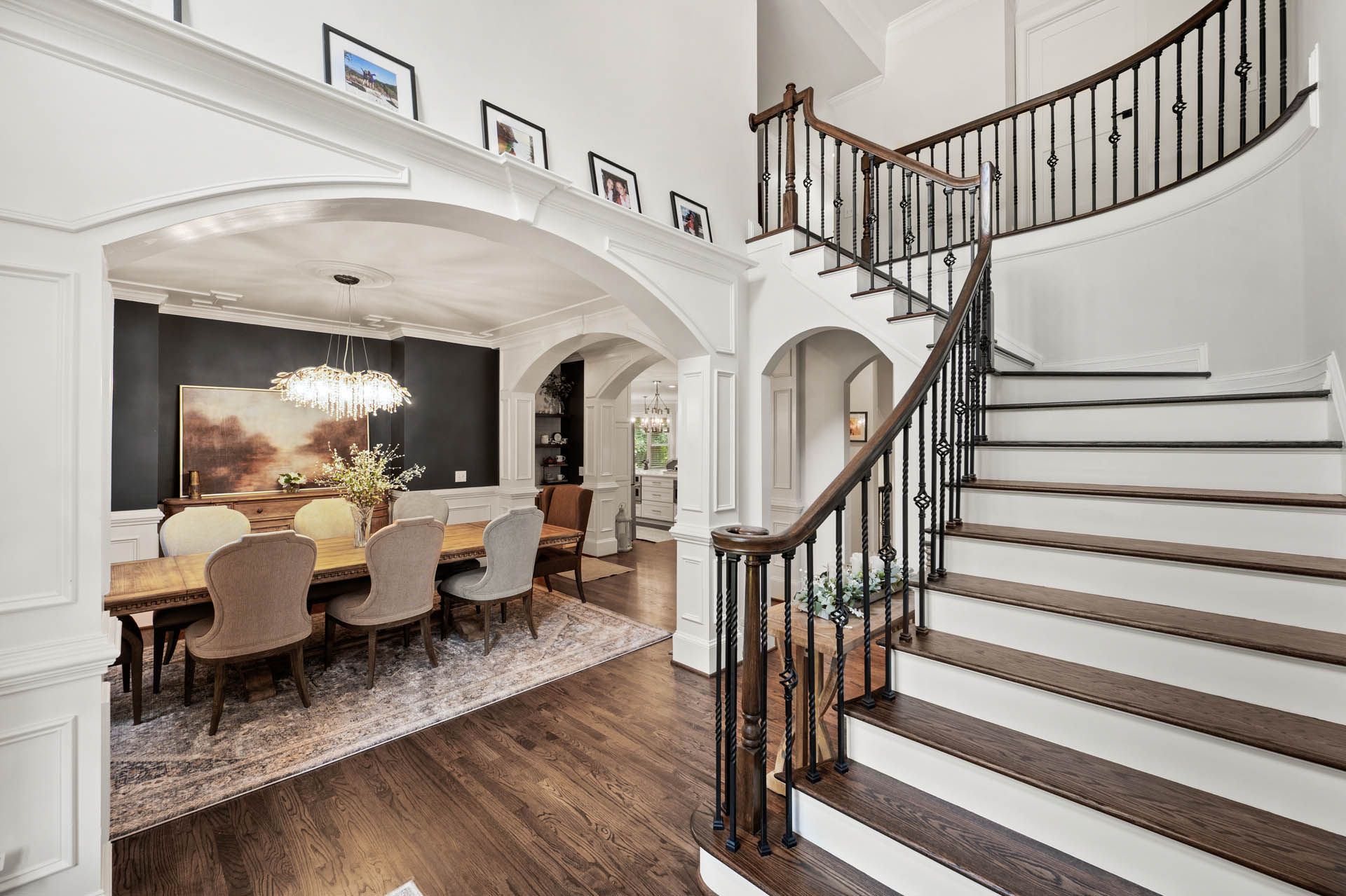 A grand entryway with a curved staircase and arched doorways leading to a dining room with a large table and chandelier.
