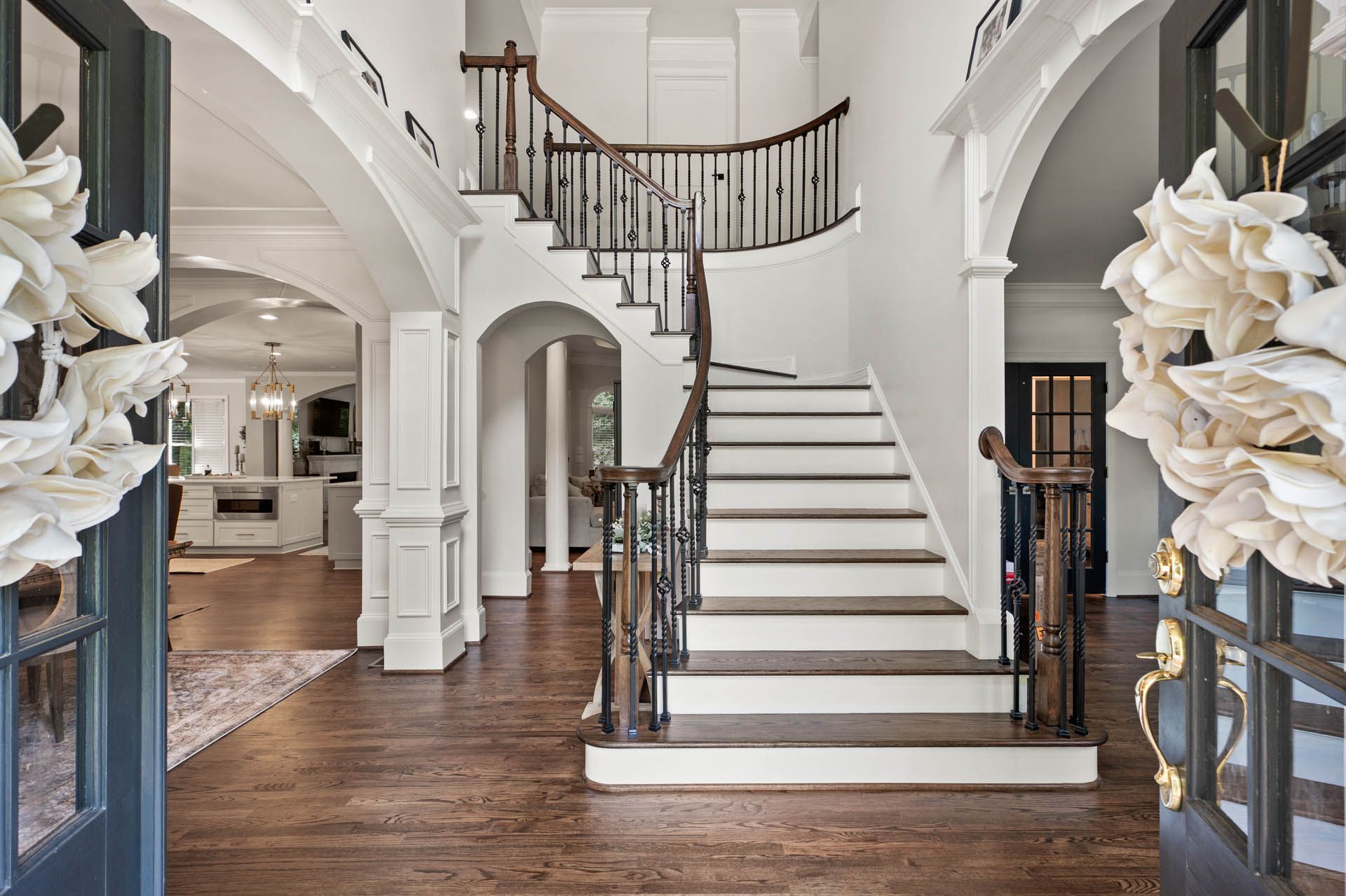 Grand foyer with a staircase, white walls, and dark wood floors. Entryway has a double door adorned with floral wreaths.