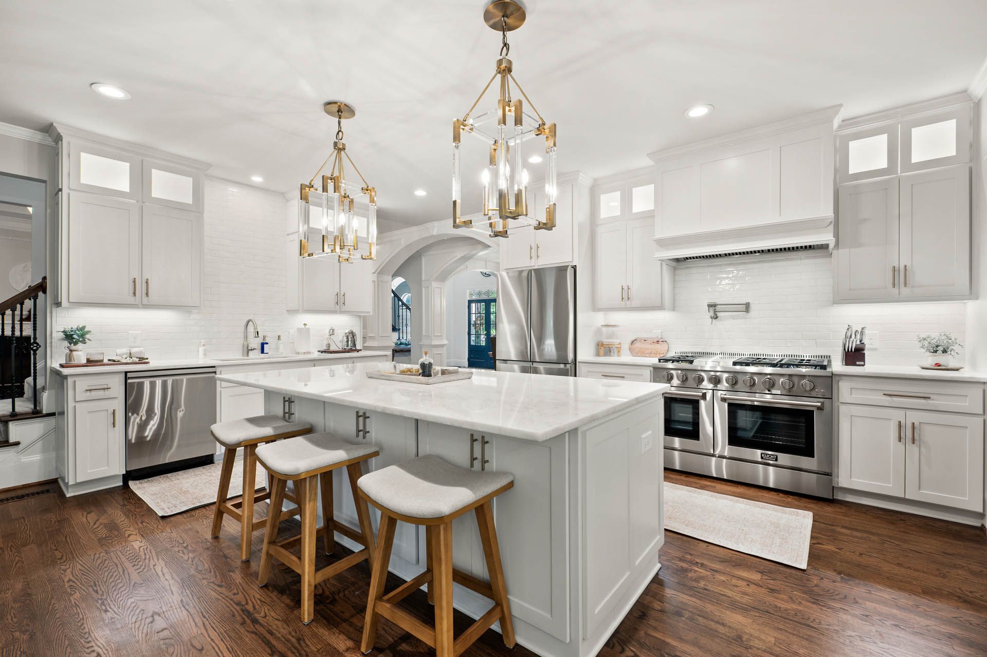 Bright, white kitchen with a large island and stainless steel appliances. Three stools sit at the island. Two gold chandeliers hang overhead.