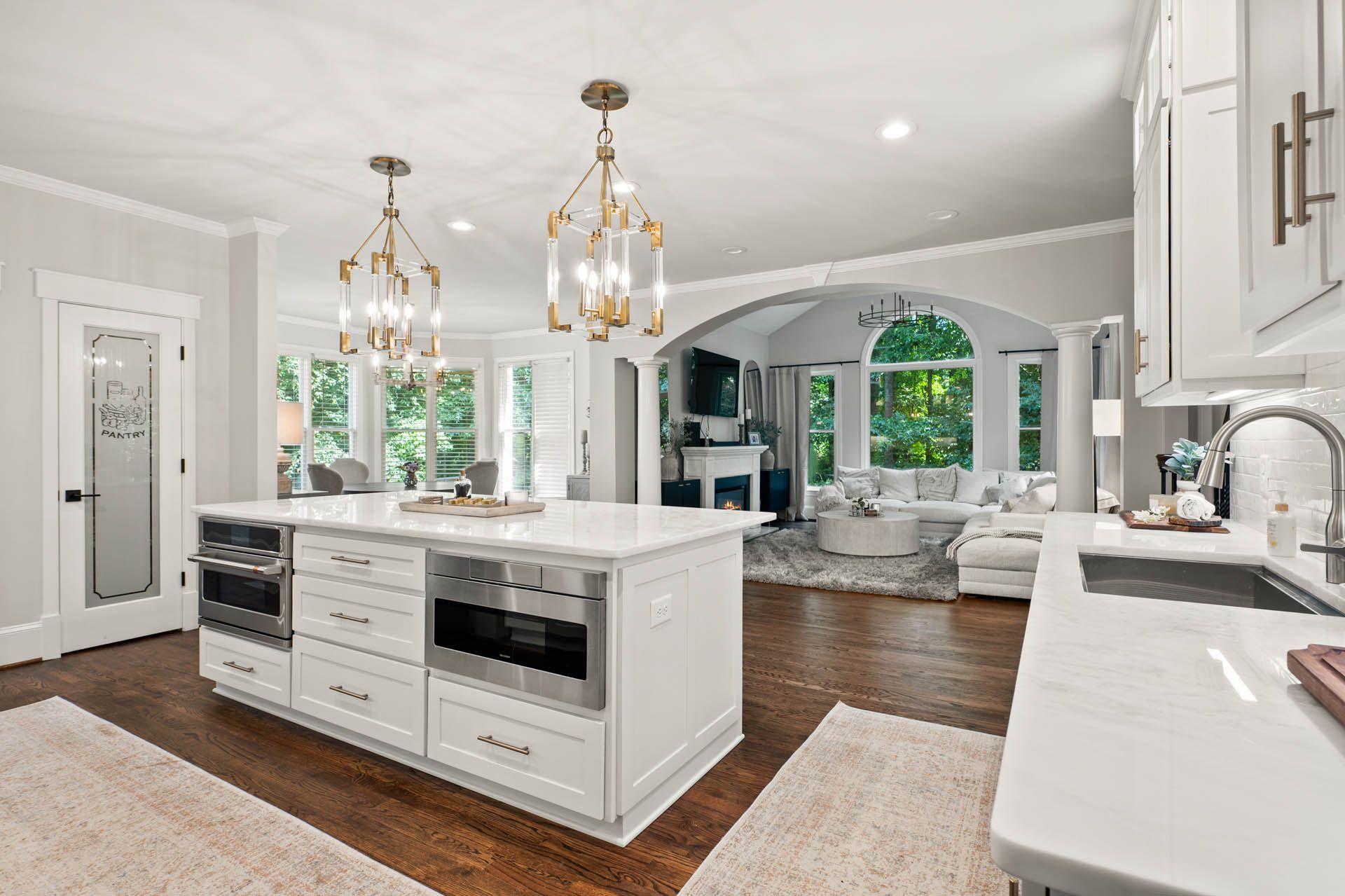 Spacious, bright kitchen with white cabinets, marble countertops, and a large island featuring built-in appliances. Open to a living area with lots of natural light.