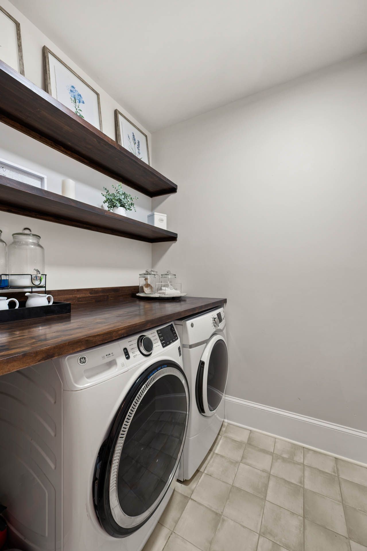 Laundry room with white washer and dryer, dark wood countertop, and open shelving with decor. Gray walls and tiled floor.