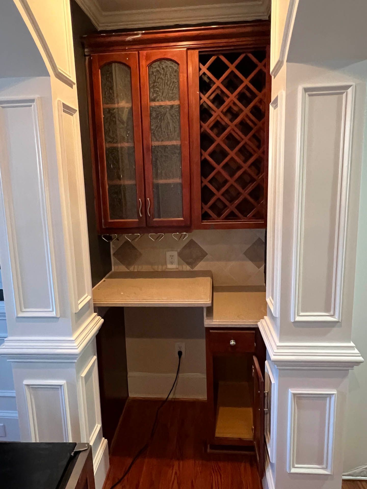 Built-in wooden bar area with wine rack, cabinet, and countertop nestled between two white pillars, viewed indoors.