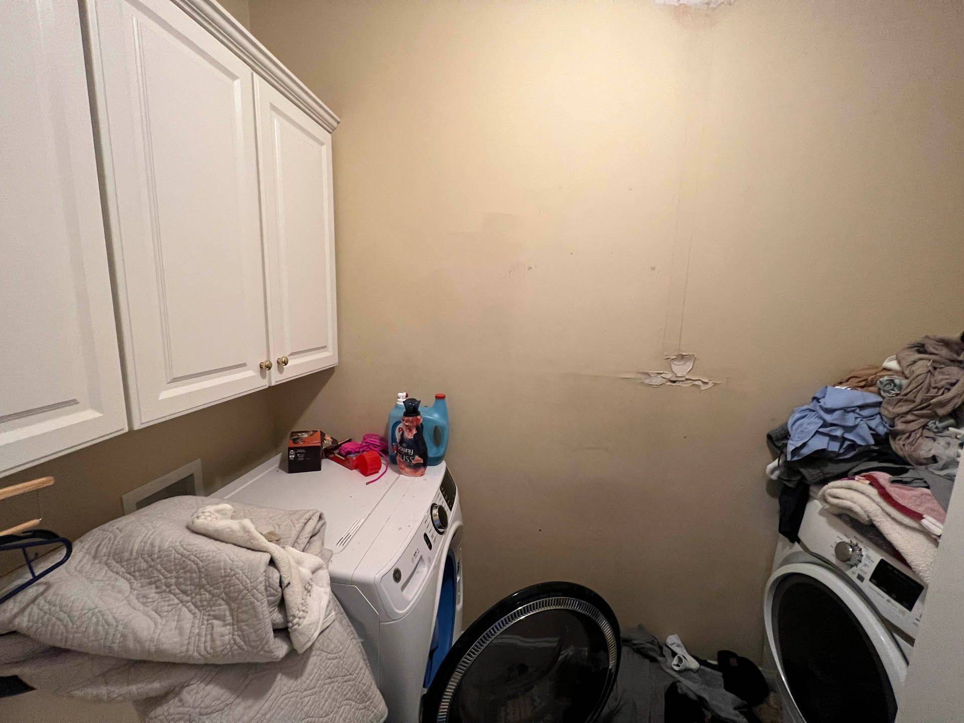 Laundry room with white cabinets, washer and dryer, and a wall with visible damage. Clothes are piled on and around the machines.