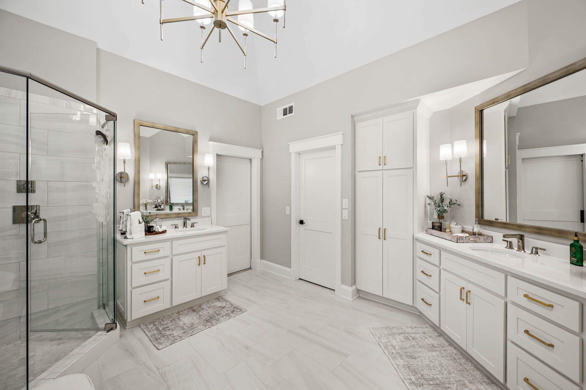 Spacious, white bathroom with marble floors and countertops, dual sinks, a glass shower, and gold fixtures.